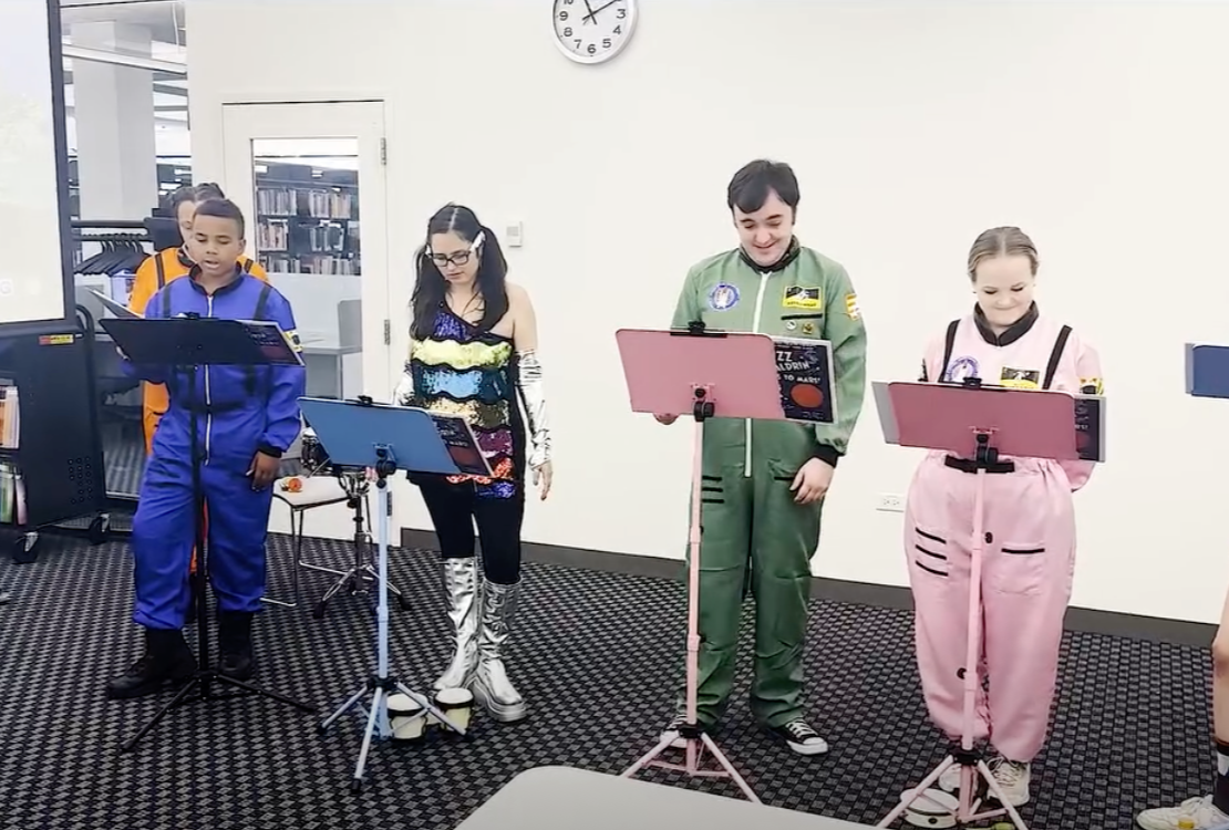 Actors performing a staged reading of a children’s theatre production at a library, standing at music stands in themed costumes.