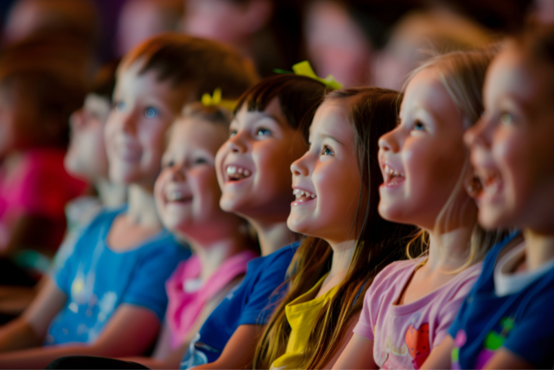 Children smiling and watching a live theatre performance with excitement and wonder.