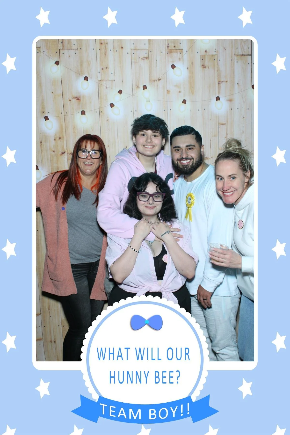A group of six people smiling and posing together at a party, with a wooden backdrop and string lights overhead, in a photo booth frame labeled 'What will our hunny bee? Team Boy!!'.