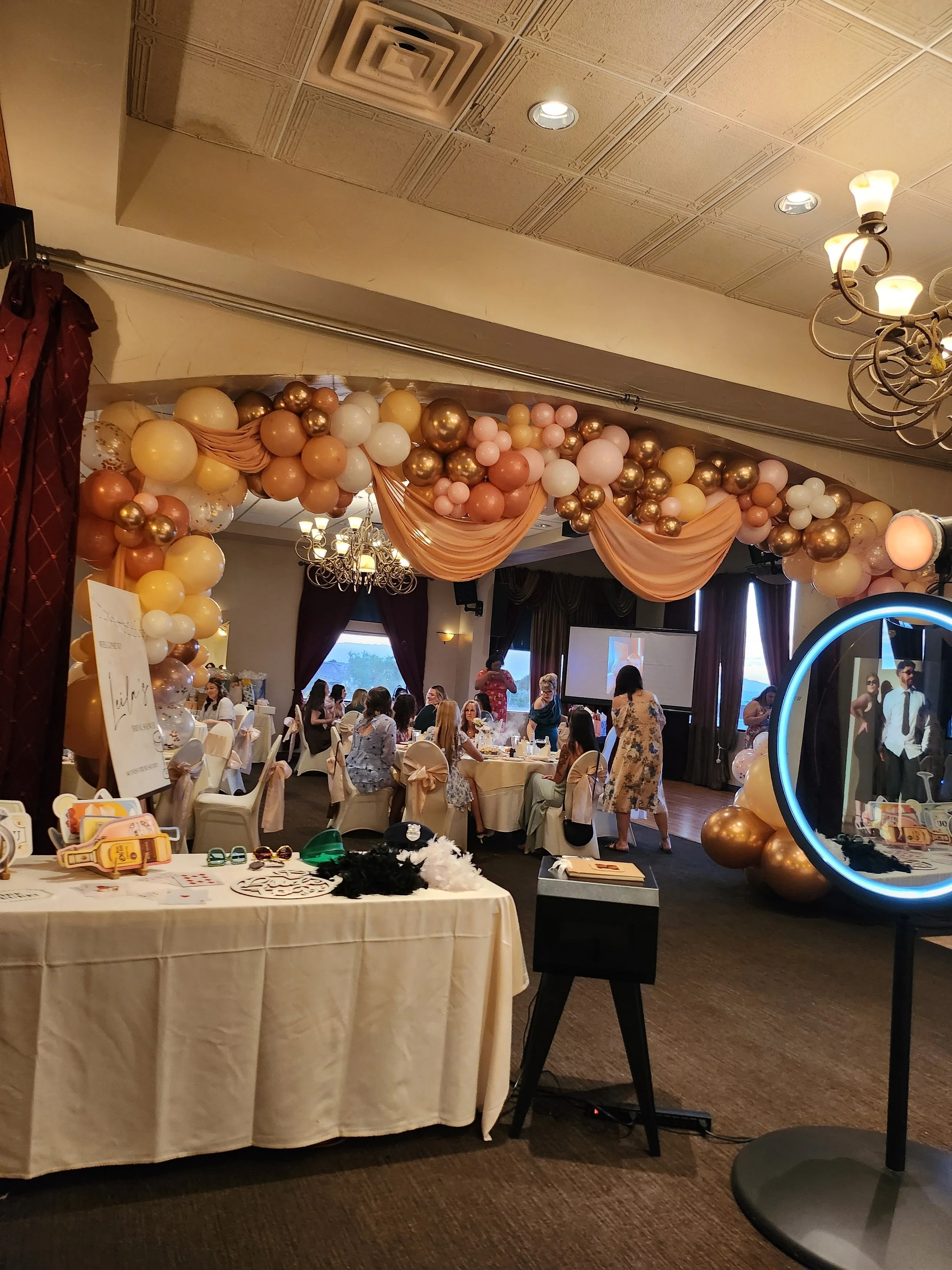 A decorated banquet hall with balloon arch, draped fabric, and chandelier, filled with guests seated at round tables for a celebration event.
