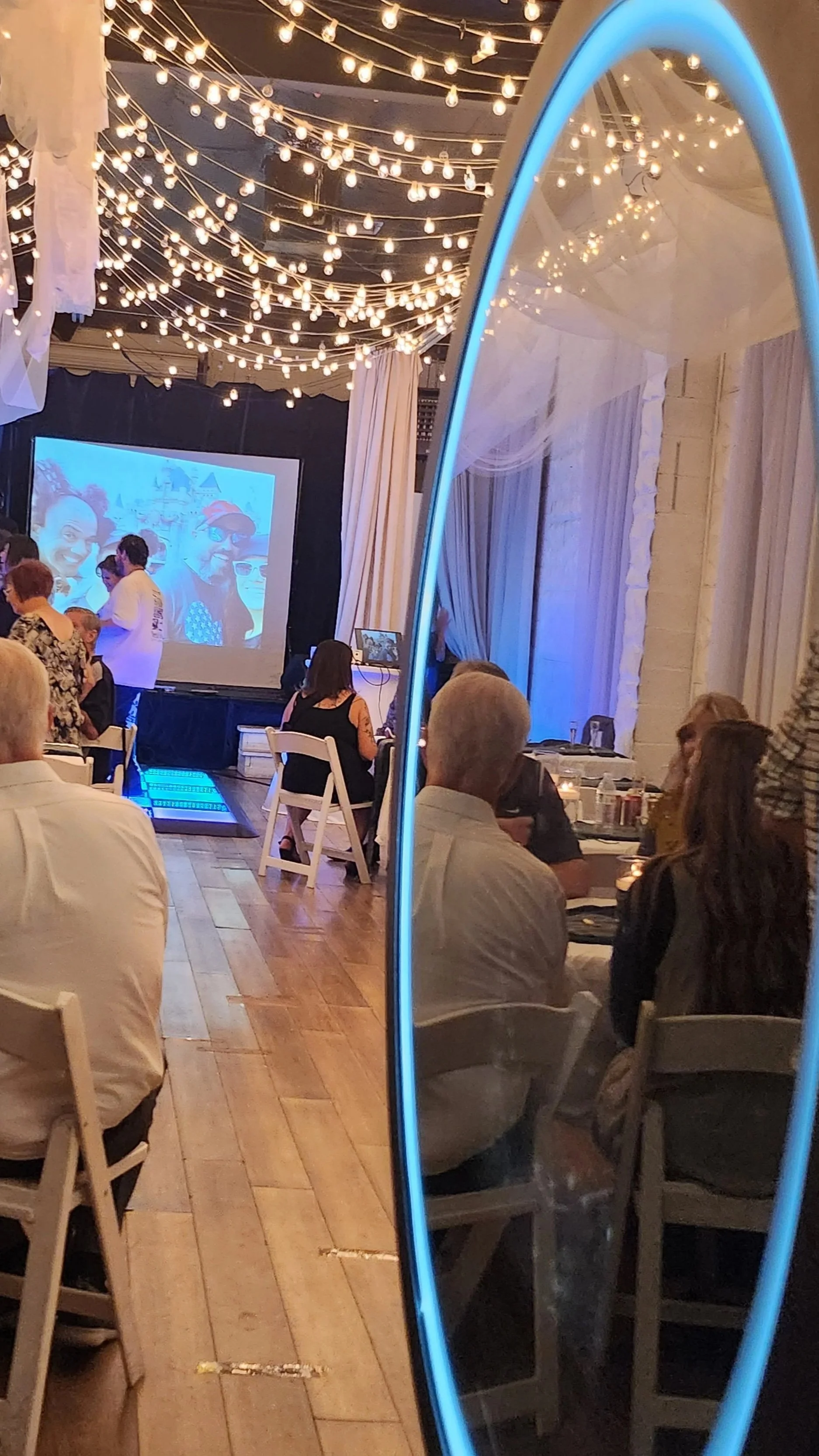 People attending a celebration or event, sitting at tables, watching a projector screen with a colorful image, decorative string lights hanging from the ceiling, and a large mirror reflecting part of the scene.
