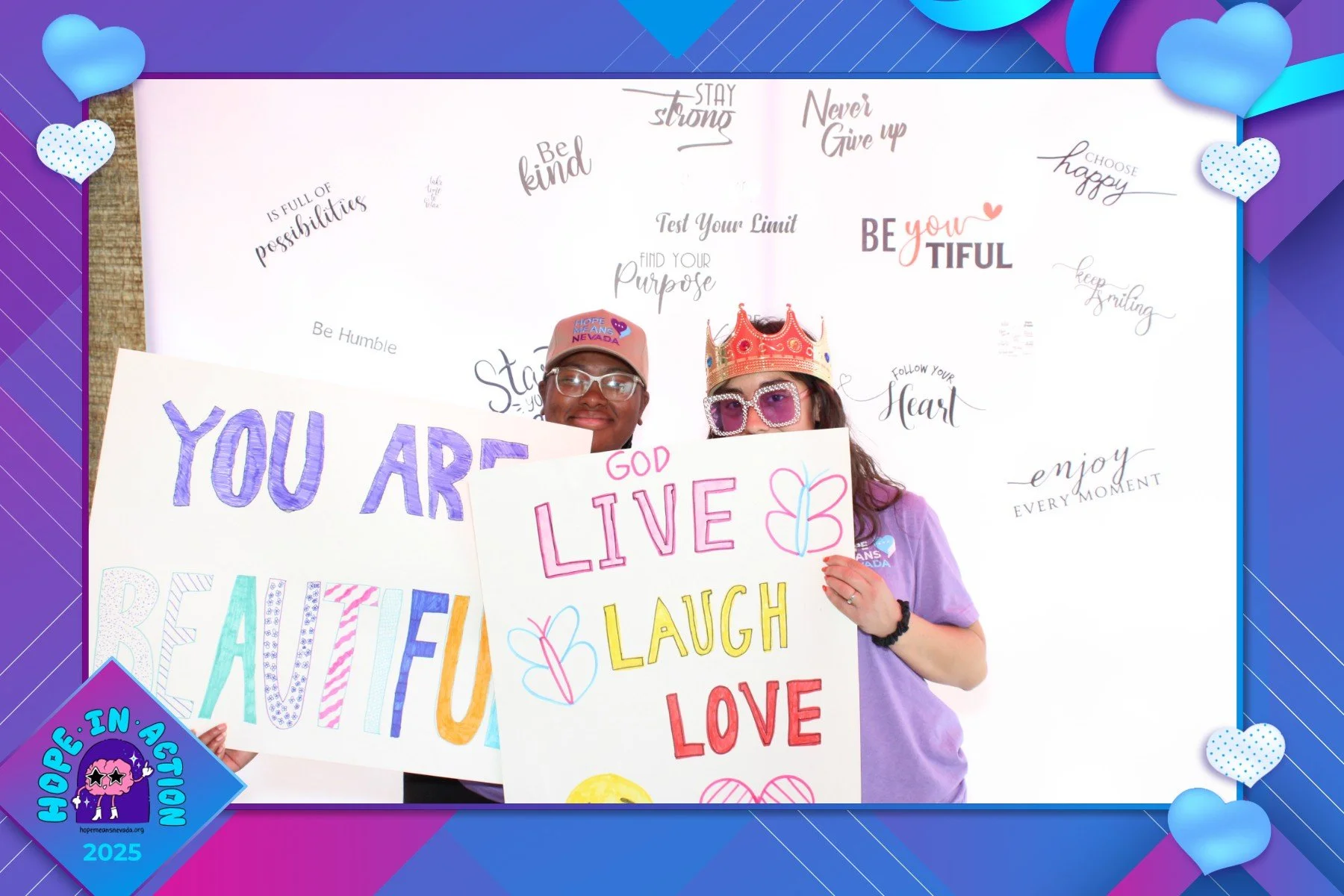 Two women smiling and holding colorful signs with positive messages, one wearing a crown and sunglasses, the other wearing a pink cap, in front of a white background decorated with inspirational quotes and heart-shaped decorations.