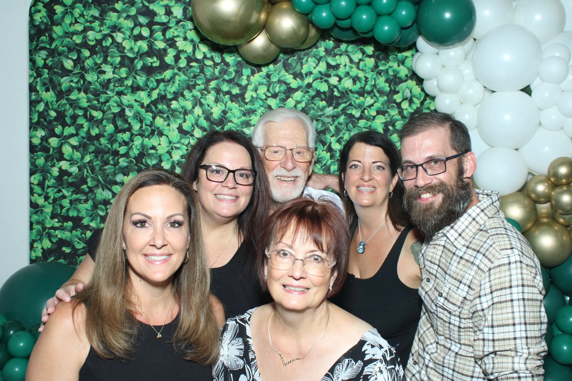 A group of seven people, six women and one man, smiling and posing together in front of a green leafy and balloon backdrop for a celebration or gathering.