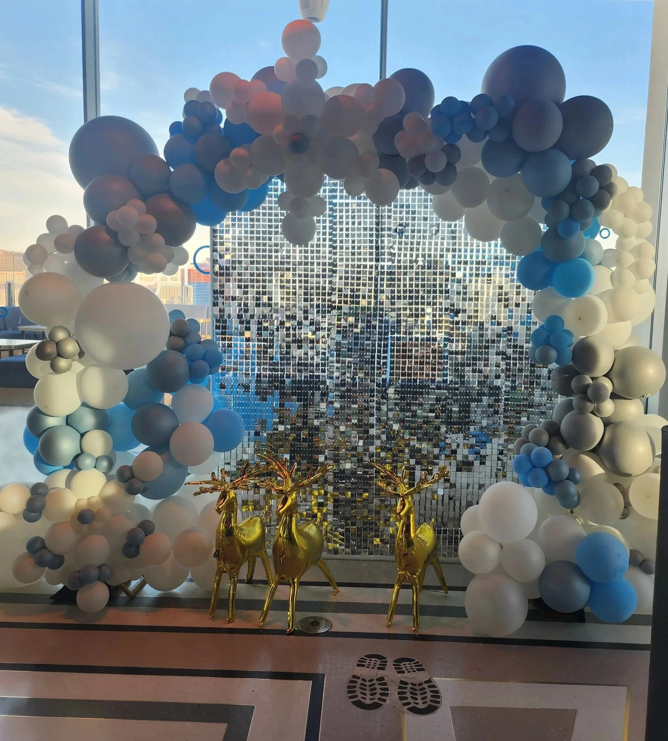 Decorative arch made of various sized white, gray, and blue balloons, with three gold reindeer statues positioned in front of a mosaic window that reflects a cityscape. The setting appears to be inside a building with large windows and a view of the city skyline.