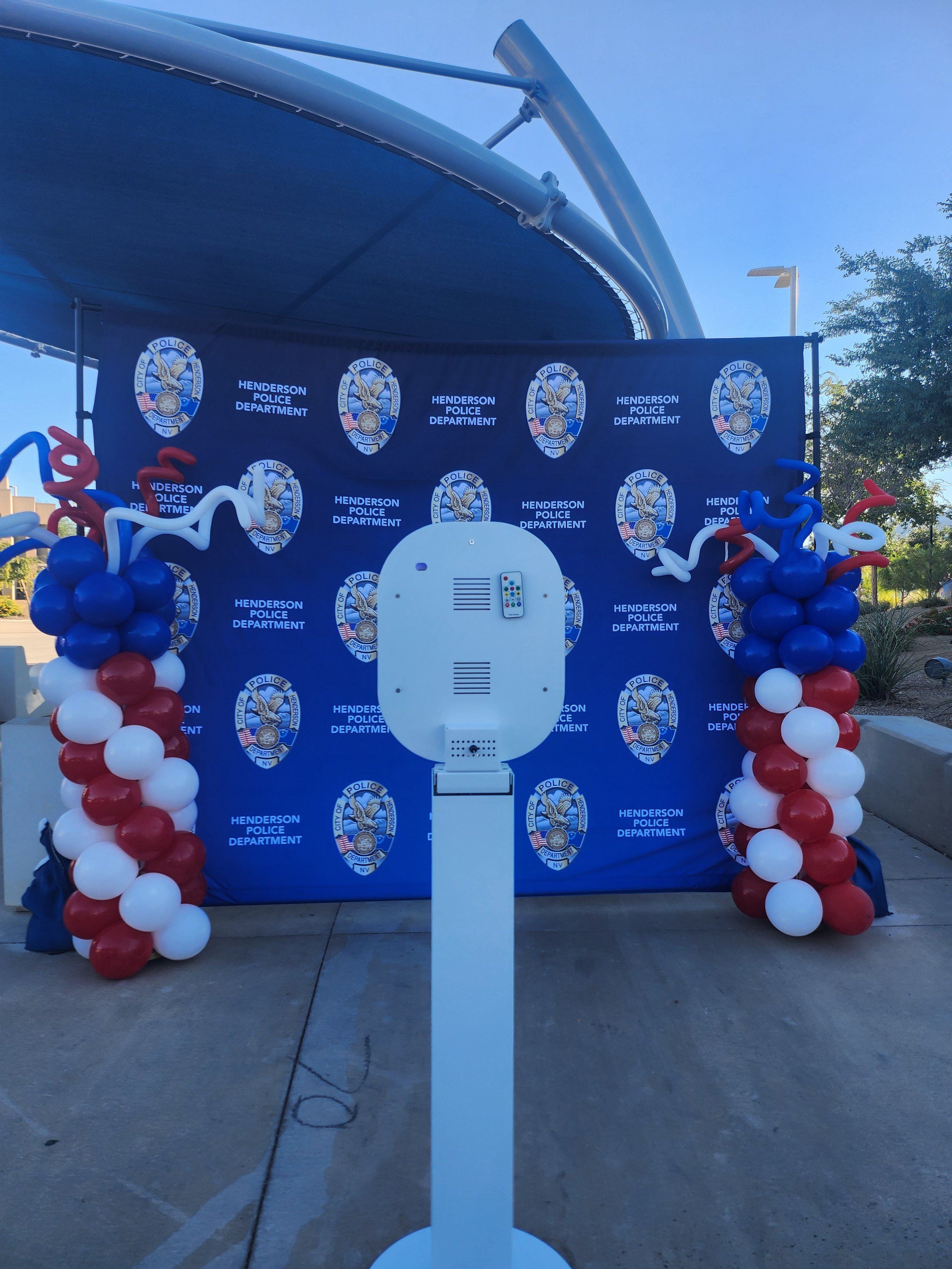 A decorated area at the Henderson Police Department with a backdrop featuring police badges and balloons in red, white, and blue, and a white police safety shield in the foreground.