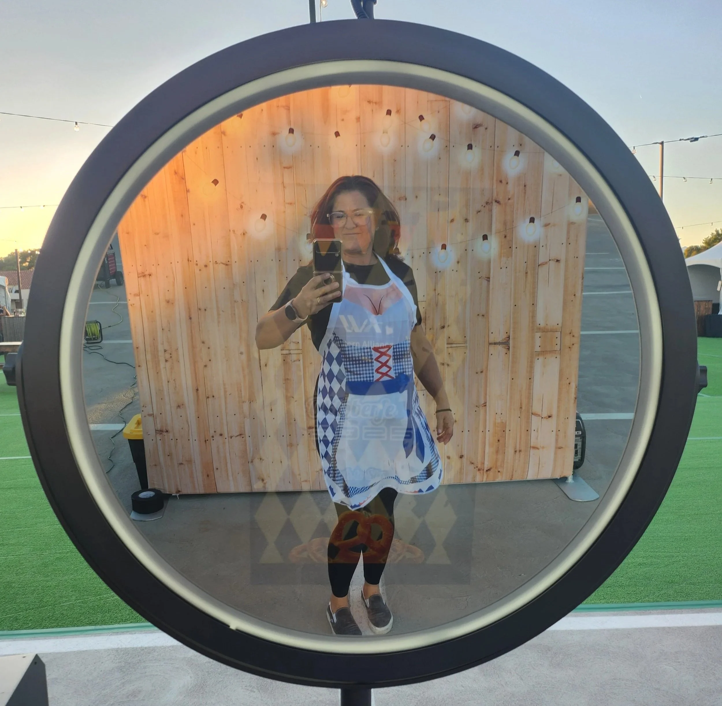 A woman in a black top and checkered apron taking a selfie in a circular mirror, with a wooden backdrop and string lights outside during sunset.