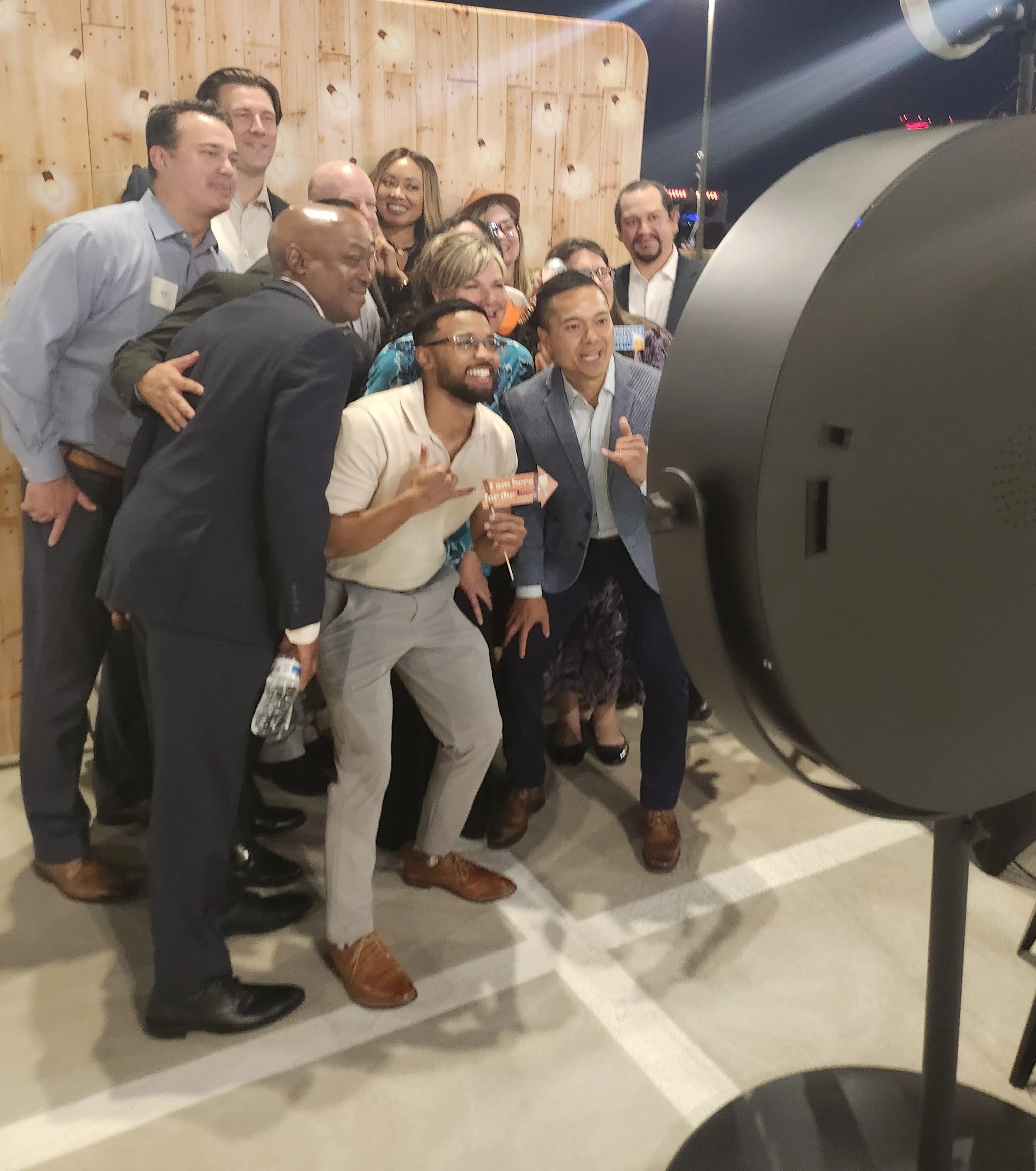 Group of people taking a photo together using a photo booth, with a large camera in the foreground and a wooden wall background.