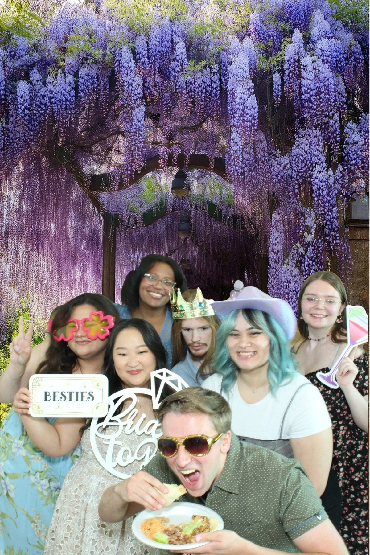 A group of seven friends celebrating at a party with a purple flower-covered backdrop, some wearing fun props like a crown, large sunglasses, and a cowboy hat, holding signs labeled 'Besties' and 'Bria To', smiling, and enjoying food.