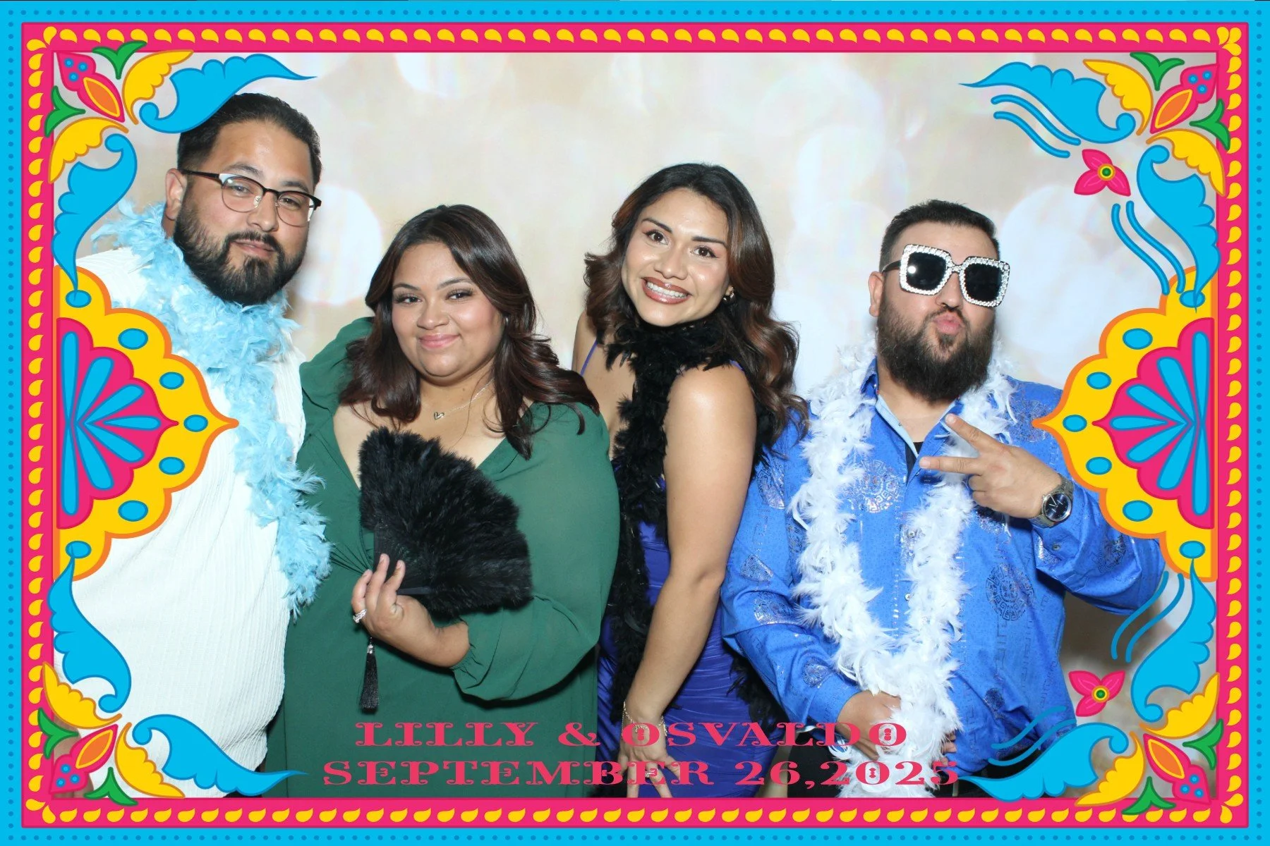 Group of five friends posing in colorful, festive photo frame with text 'Lilly & Osvaldo September 26, 2025' at the bottom, some wearing feather boas and sunglasses, celebrating at an event.