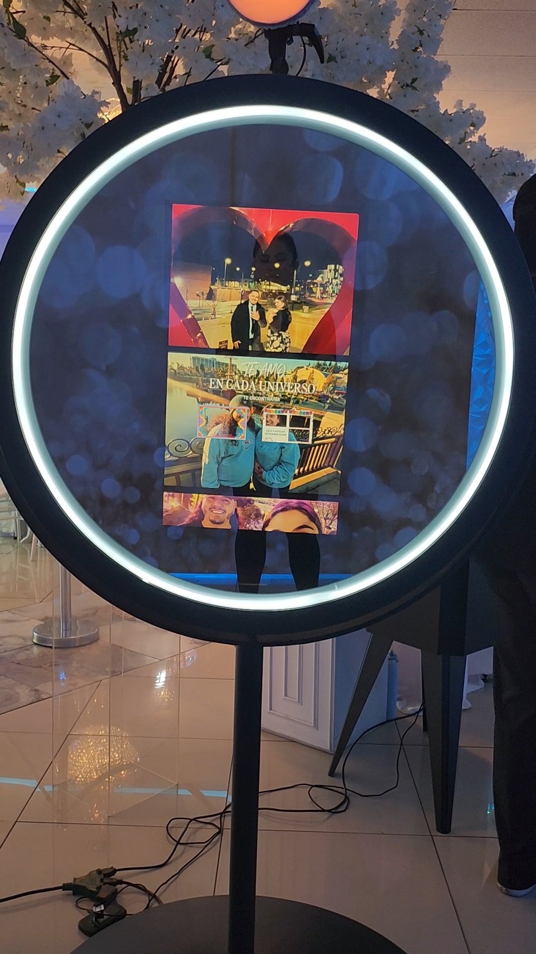 A circular photo booth with a backlit border, displaying a collage of photographs of various couples and groups, including a couple with a large heart-shaped frame, a couple on a city bridge, and a smiling man with a woman. The setting appears to be indoors with tiled flooring, white walls, and decorative lighting.