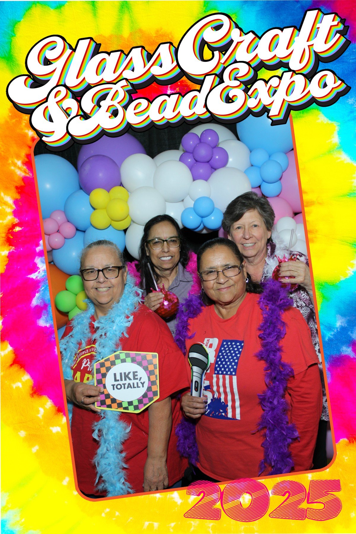 A group of four older women smiling at a Glam Craft & Bead Expo photo booth with colorful balloon decorations in the background. They are wearing festive accessories like feather boas and holding fun signs and props, celebrating the year 2025.