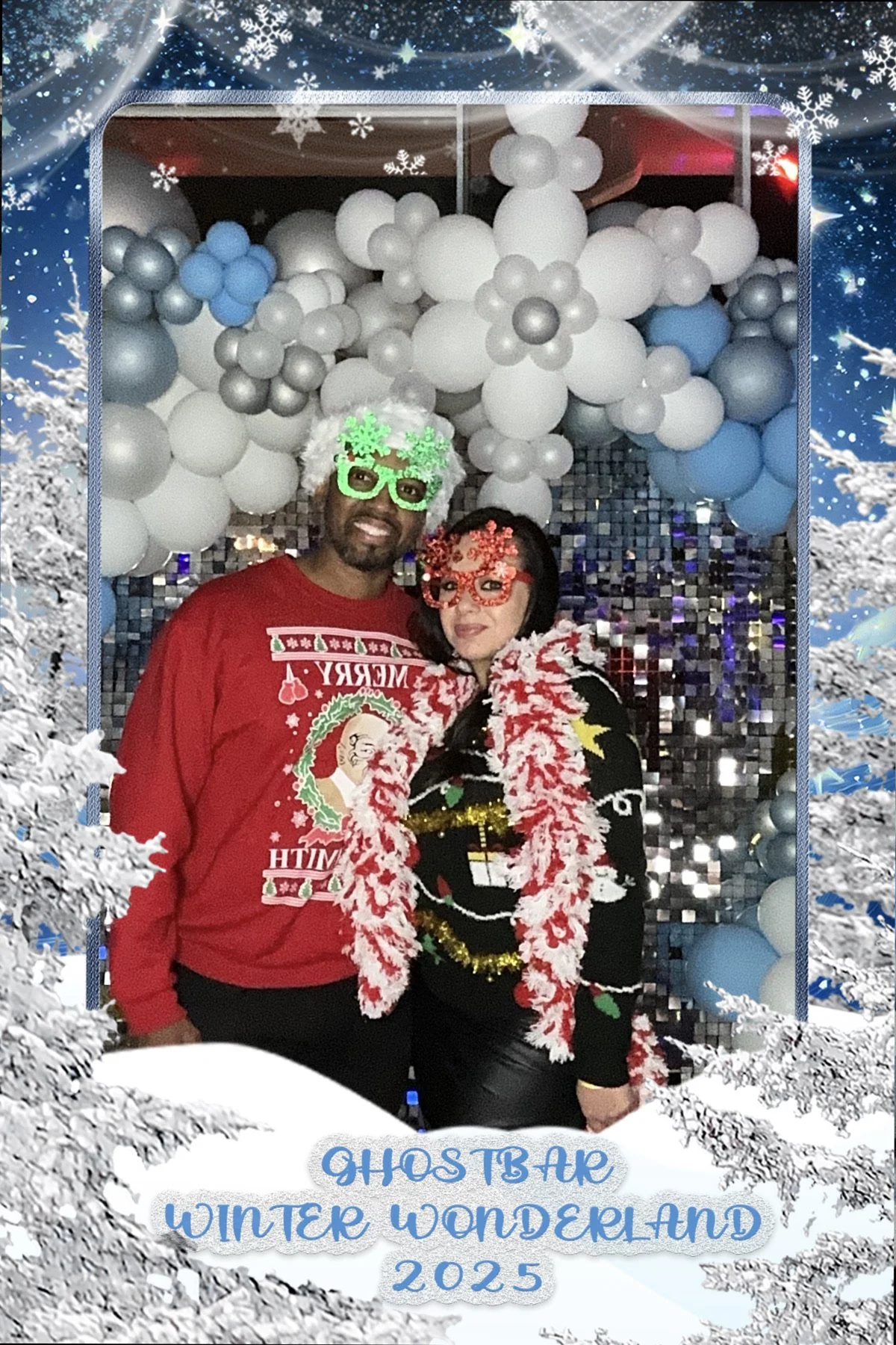 Two people at a winter-themed party, dressed in Christmas costumes with festive accessories, standing in front of a balloon backdrop with snowflake decorations. The photo frame has a snowy border with the text 'GHOSTBAR WINTER WONDERLAND 2025' at the