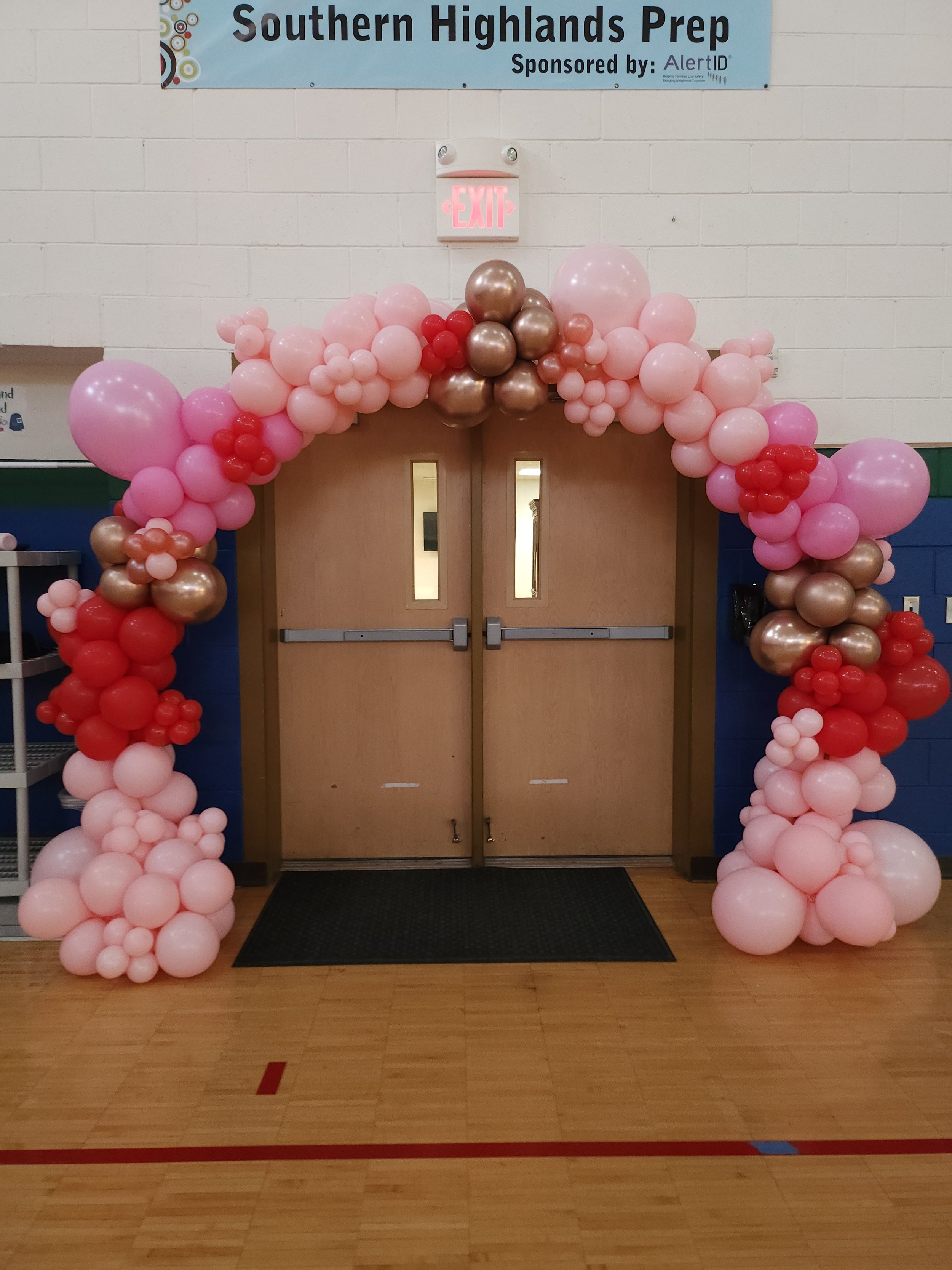 Balloon arch decorated with pink, red, gold, and peach balloons over closed double doors at Southern Highlands Prep school.