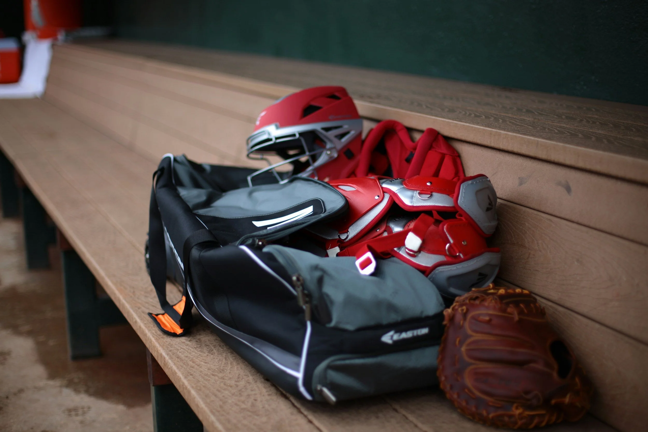 Baseball gear on a wooden park bench including a red helmet, red and gray shin guards, a black and gray bag, a brown baseball glove, and a gray and black backpack.