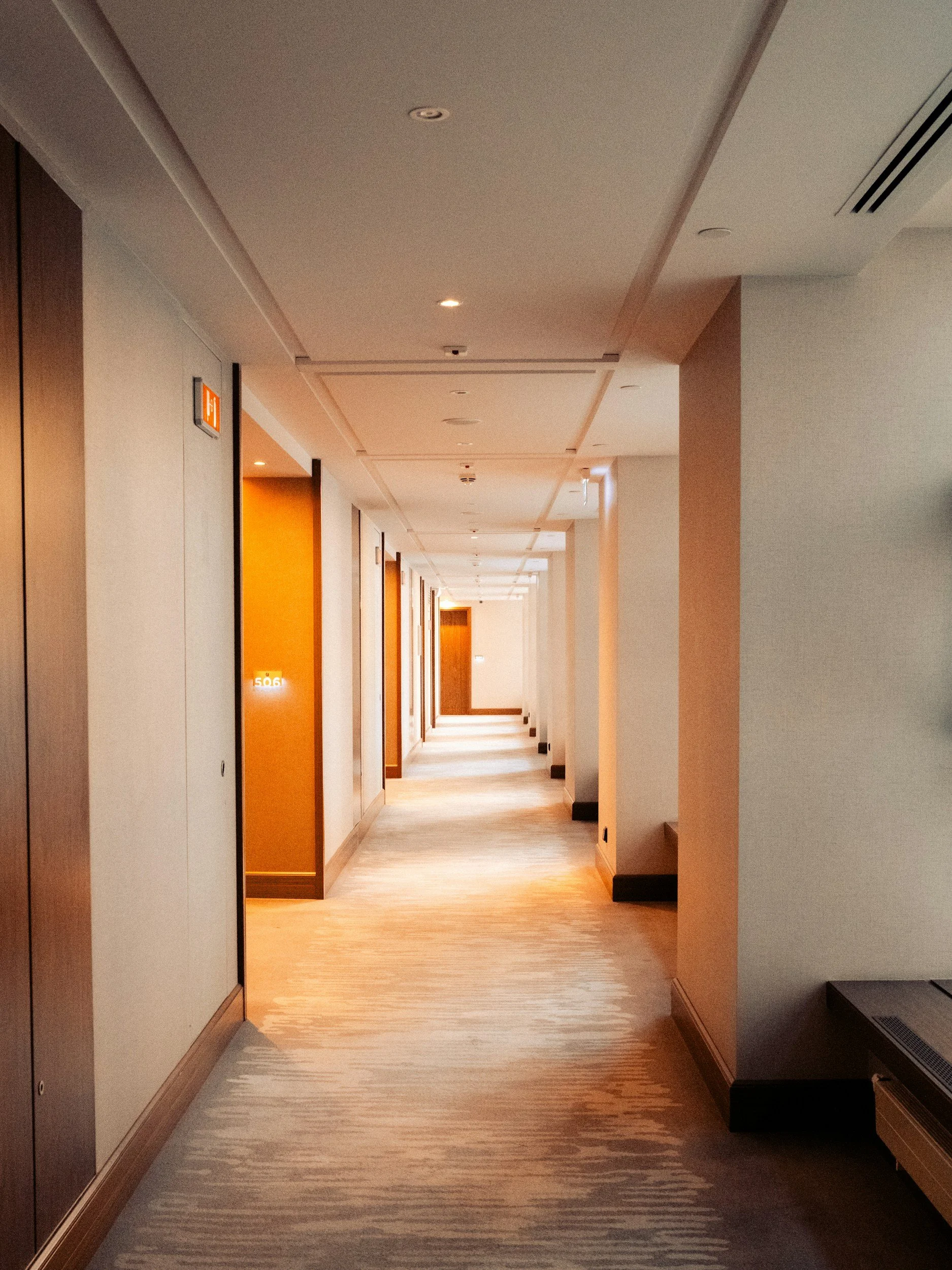 Hotel hallway with multiple closed wooden doors, some with illuminated room numbers, carpeted floor, and ceiling lights.