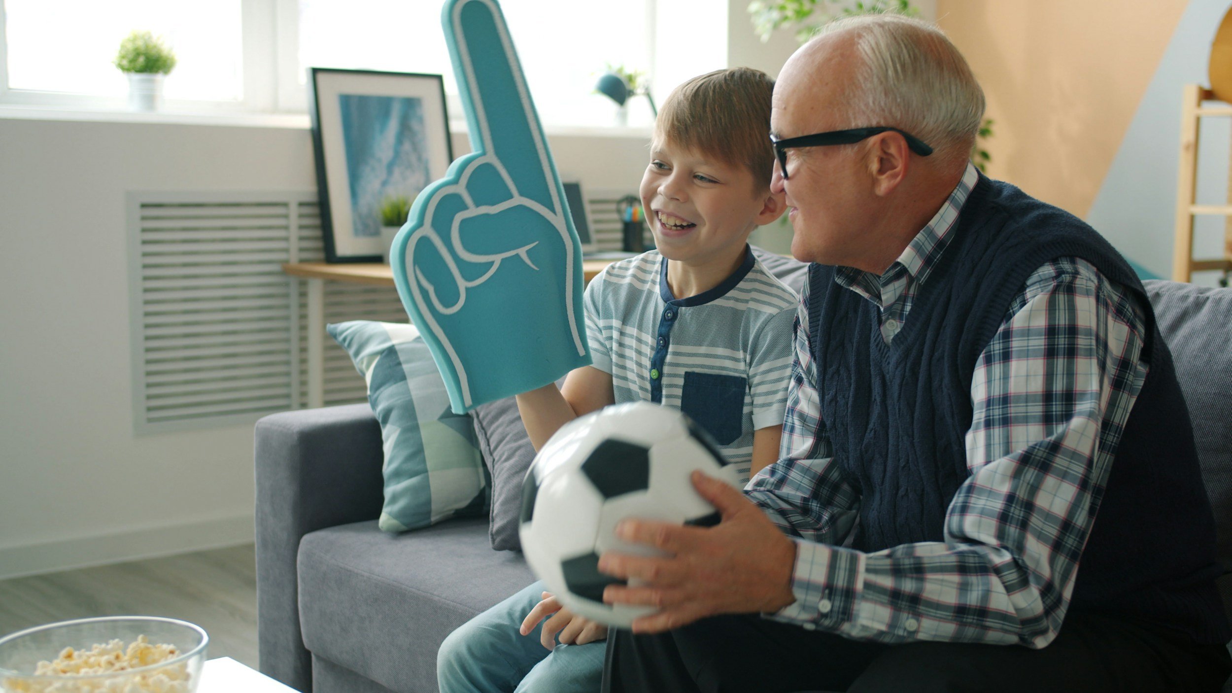 A grandfather and young boy sitting on a gray sofa, watching a soccer game on TV. The boy is holding a foam finger and smiling, while the grandfather holds a soccer ball. The room has a window with potted plants and framed pictures.