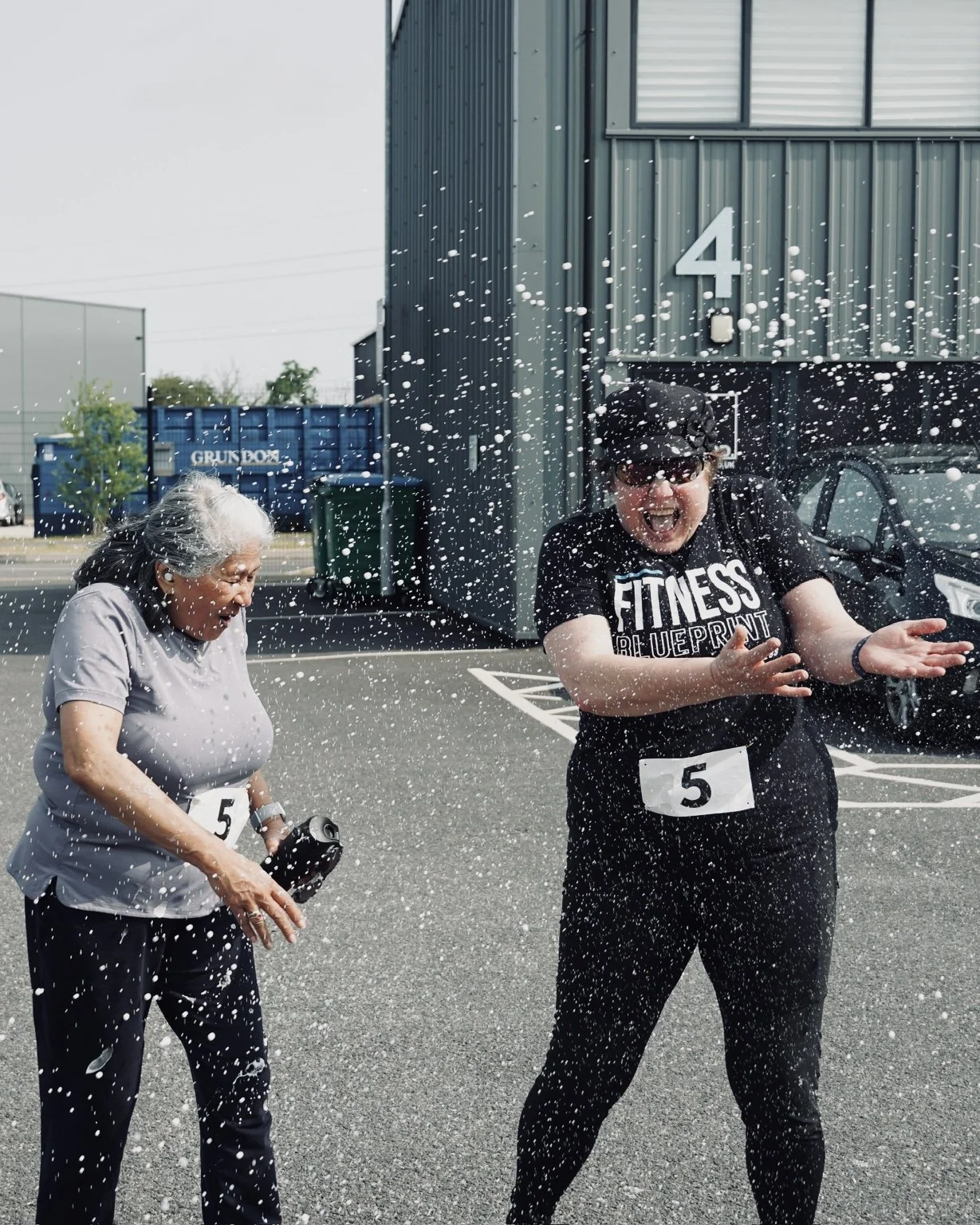Two women celebrating after a race, one older and one younger, standing in a parking lot with a building in the background. They are covered in white foam or spray, smiling and enjoying the moment.