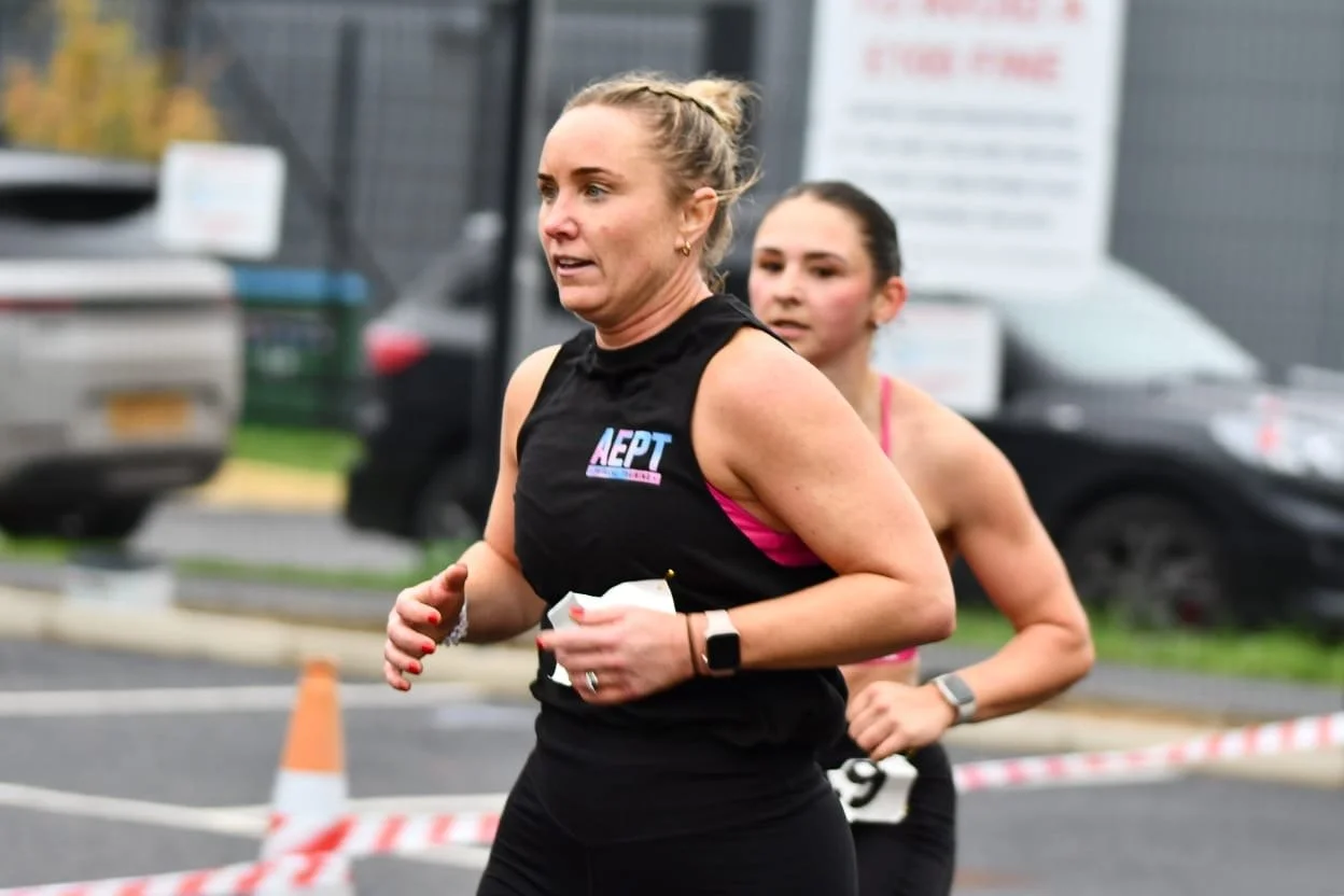 Two women running in a race, with one woman in the foreground wearing a black sleeveless top and the woman in the background wearing a pink sports bra. They are running outdoors on a roadway with parked cars and traffic cones.