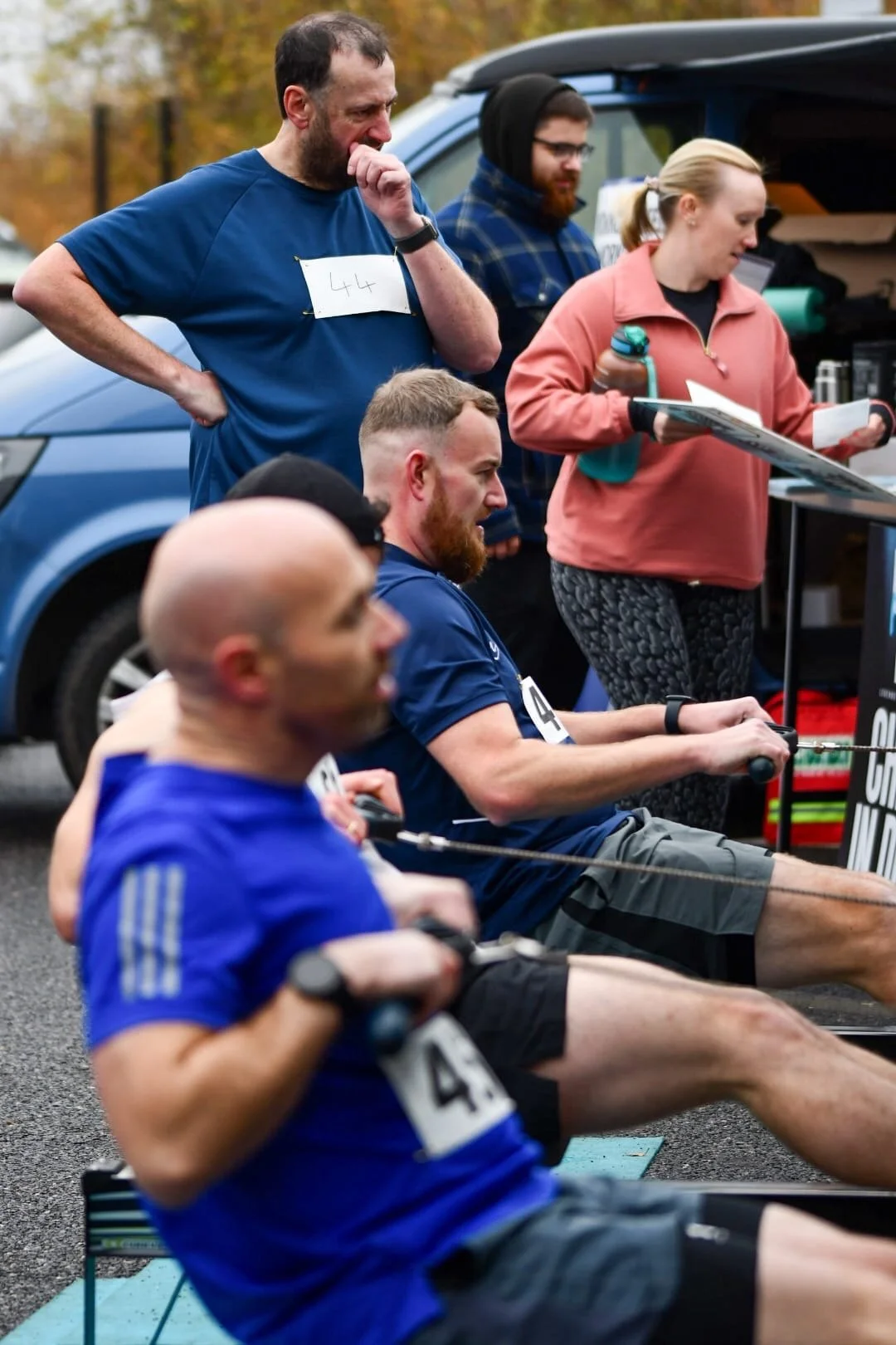 People participating in a rowing competition outdoors, with two men sitting in rowing machines in the foreground and a man and woman standing behind them, observing and taking notes.