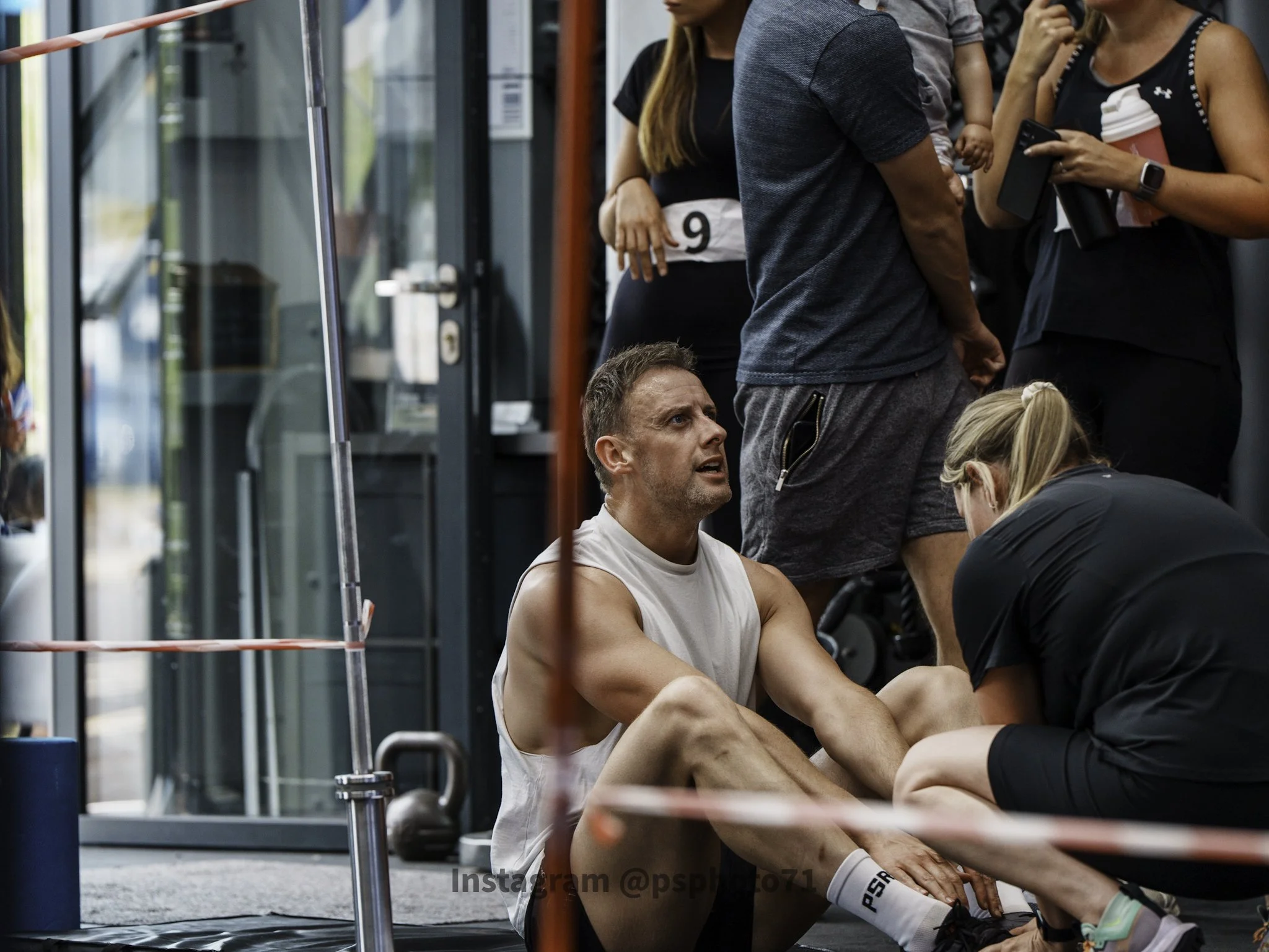 A man wearing a sleeveless shirt and shorts is sitting on the floor of a gym, holding his leg, with a woman kneeling in front of him examining his ankle. Several people surround them, some standing and talking, inside a gym or fitness center.