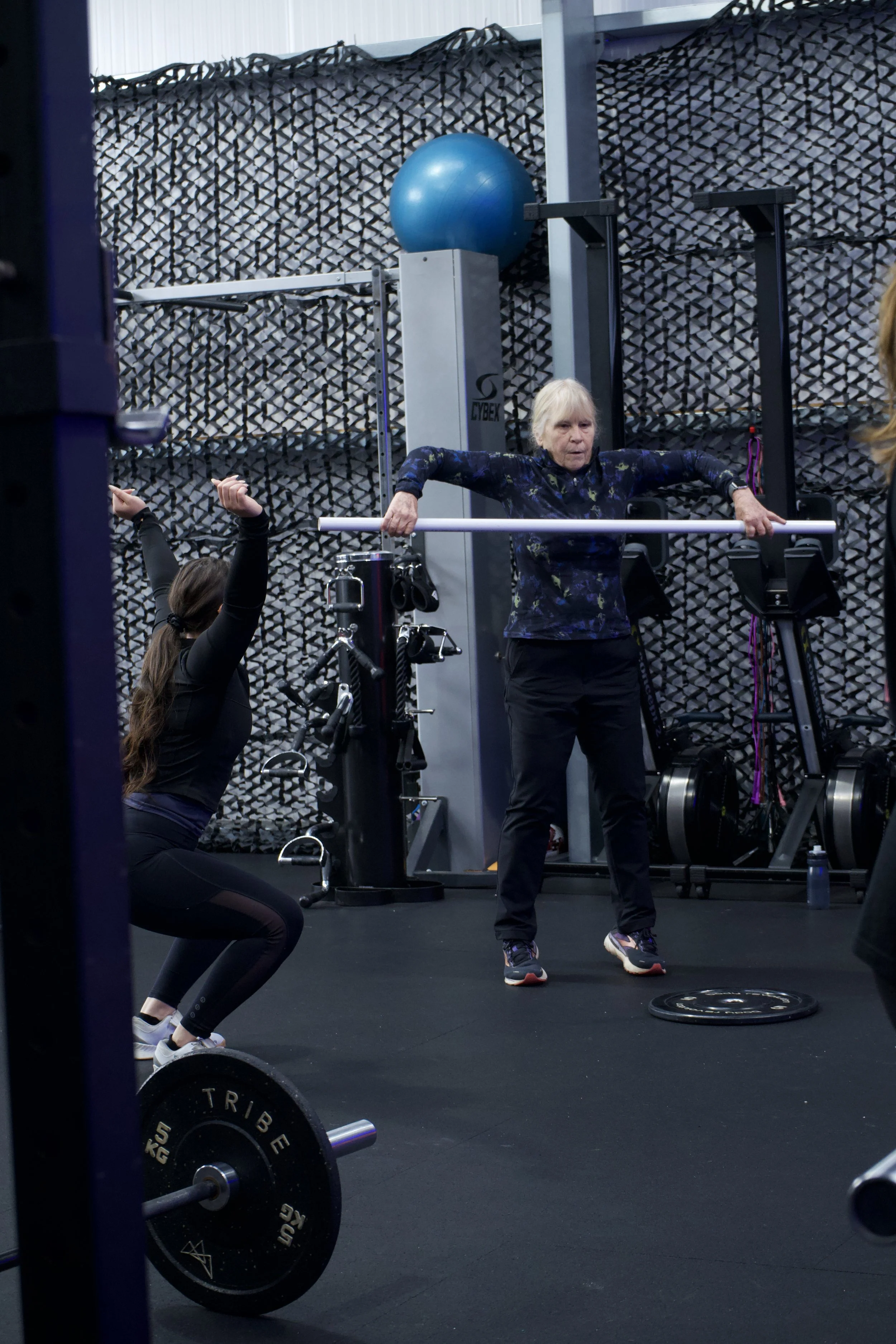 An elderly woman practicing weightlifting in a gym, with a trainer assisting her.