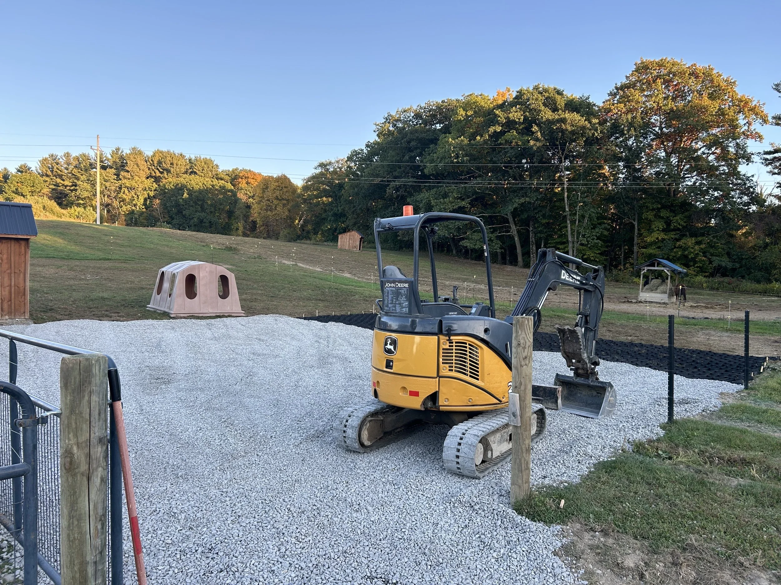 A small john deere excavator on a gravel field with a wooden post, a pink play structure, fenced area, small shed, and trees with fall foliage under a clear blue sky.
