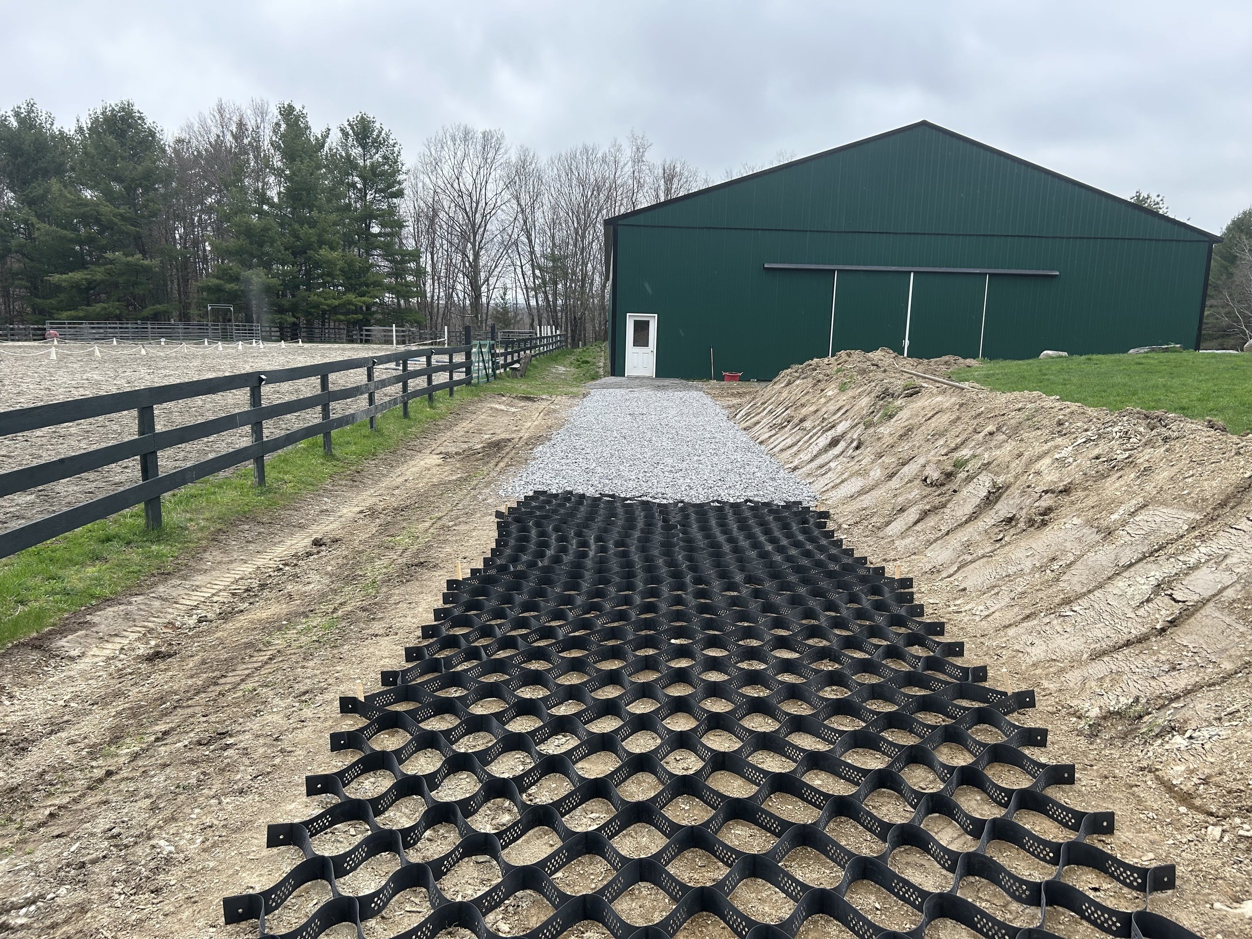 View of a farm or equestrian area with a dirt path under construction, gravel and paving mats, a green barn, trees in the background, and cloudy sky.