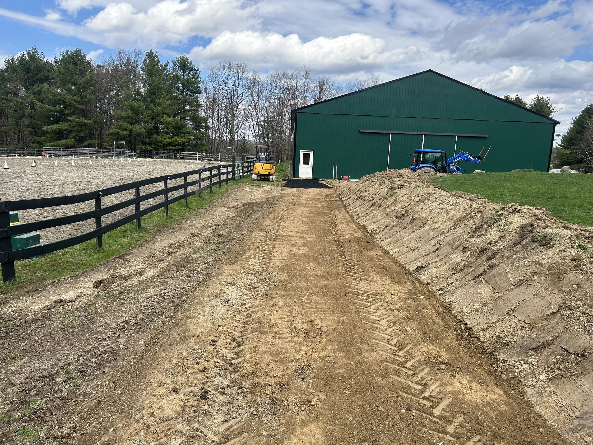 A dirt pathway beside a fenced area for horses, with a large green barn, a small white door, a front loader, and a tractor against the barn. Trees and cloudy sky are in the background.