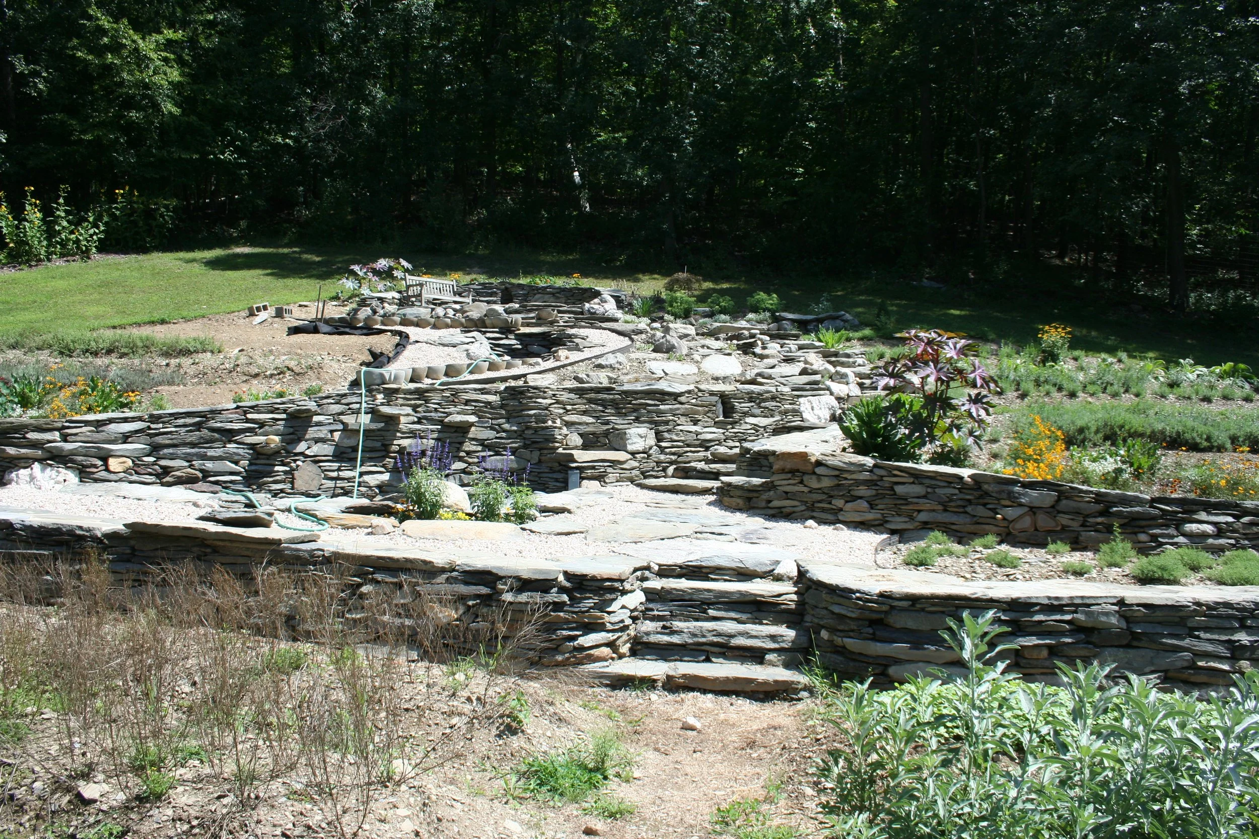 A landscaped garden with stone retaining walls, steps, and various flowering plants, bordered by a grass lawn and surrounded by trees.