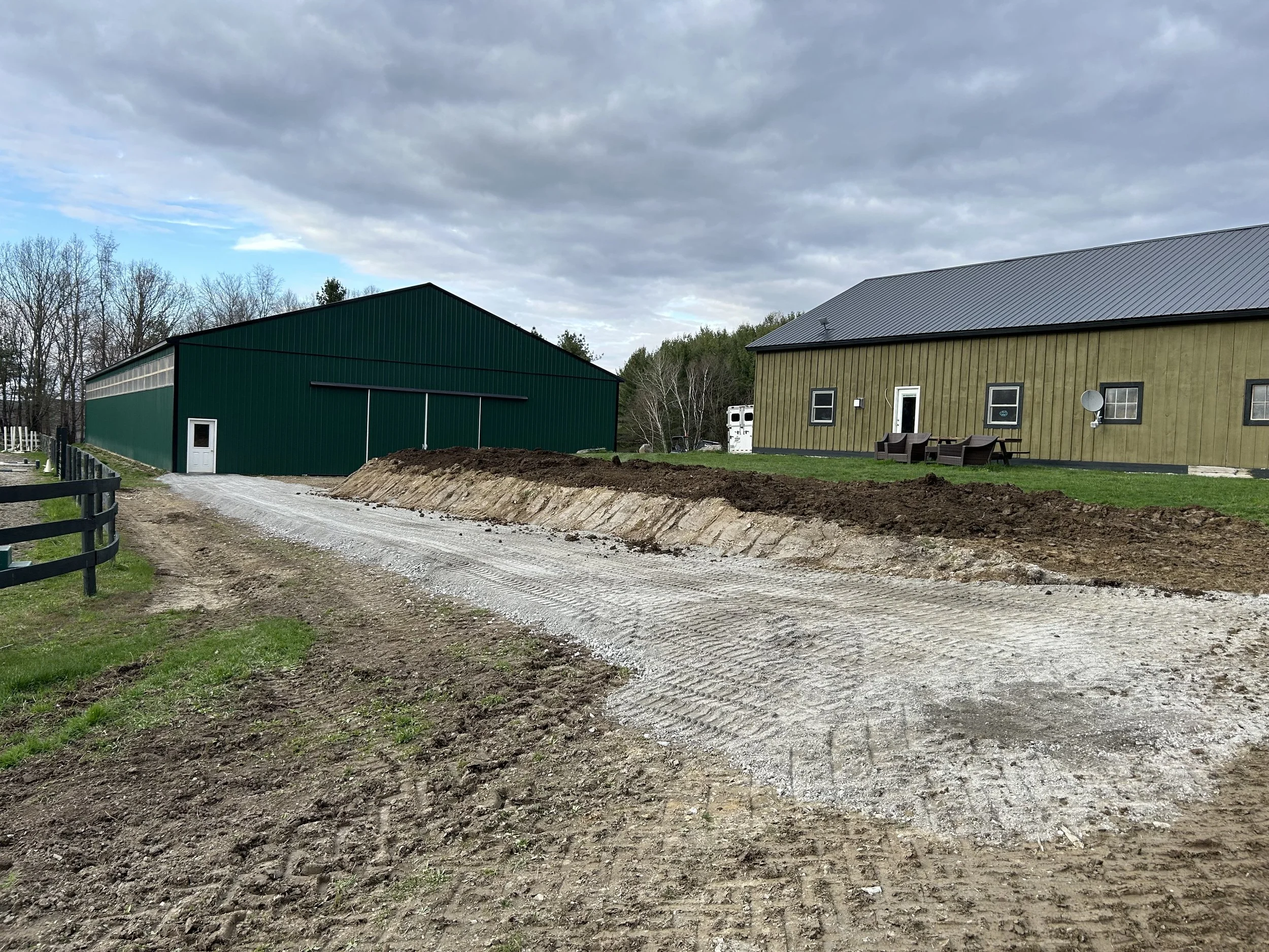 A gravel driveway leading to a green barn and a yellow house with outdoor furniture and satellite dish.