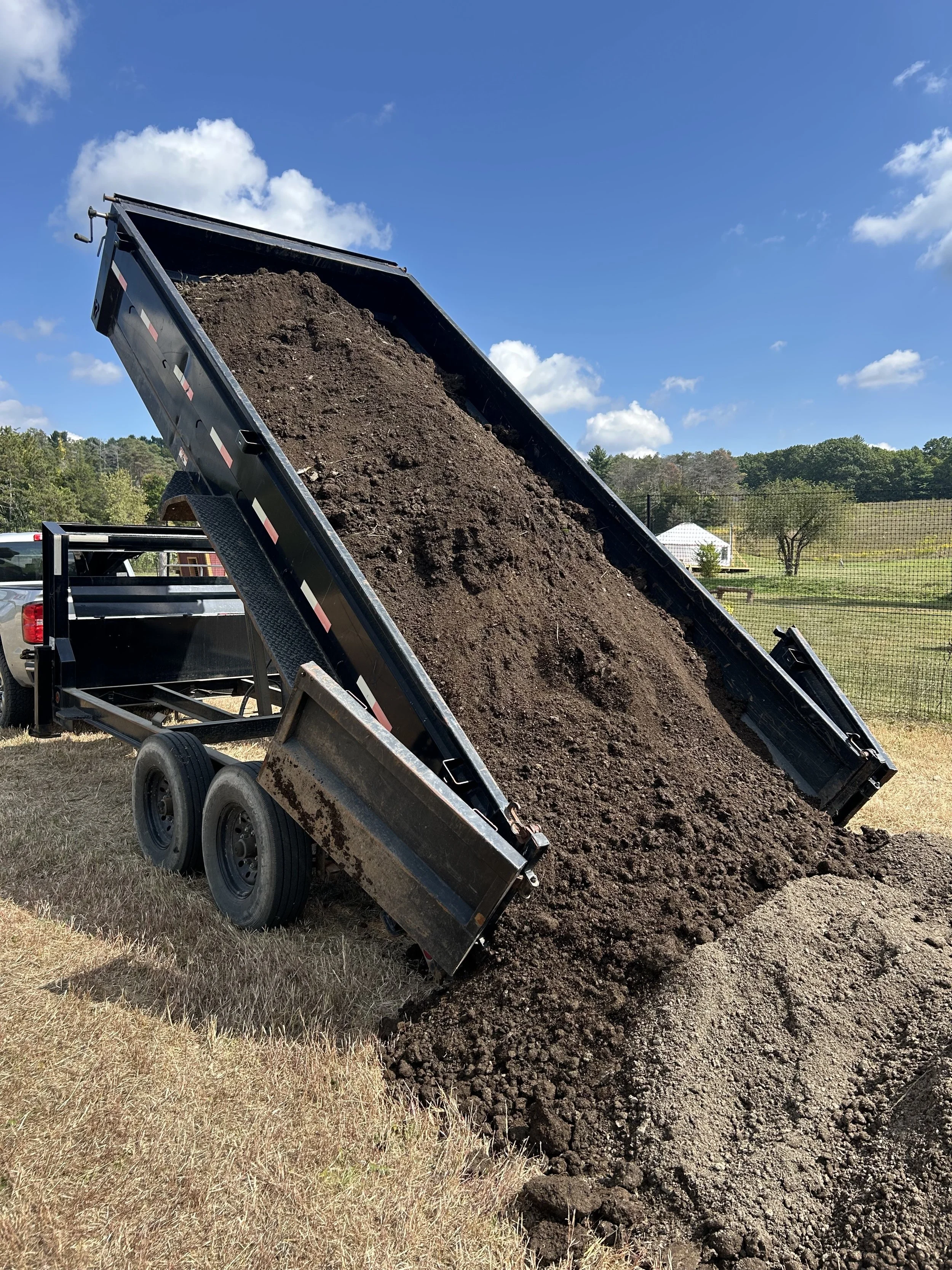 A dump truck unloading a large amount of dirt or soil onto a grassy area outdoors, with a background of trees and a partly cloudy sky.