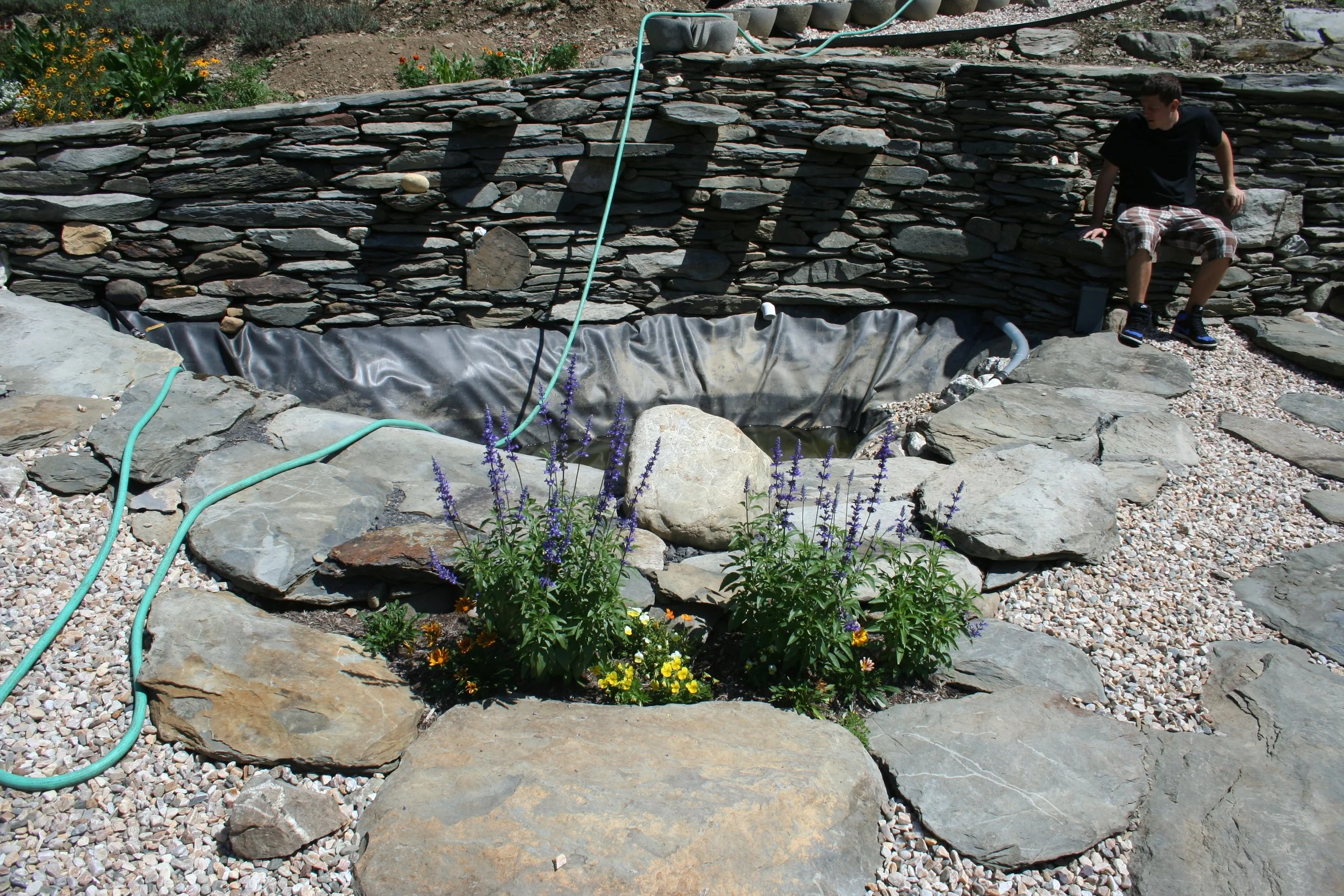 A small garden pond lined with black plastic, surrounded by large flat stones and decorative rocks, with flowering plants in front and a stone wall in the background. A person is sitting on the rocks to the right of the pond.