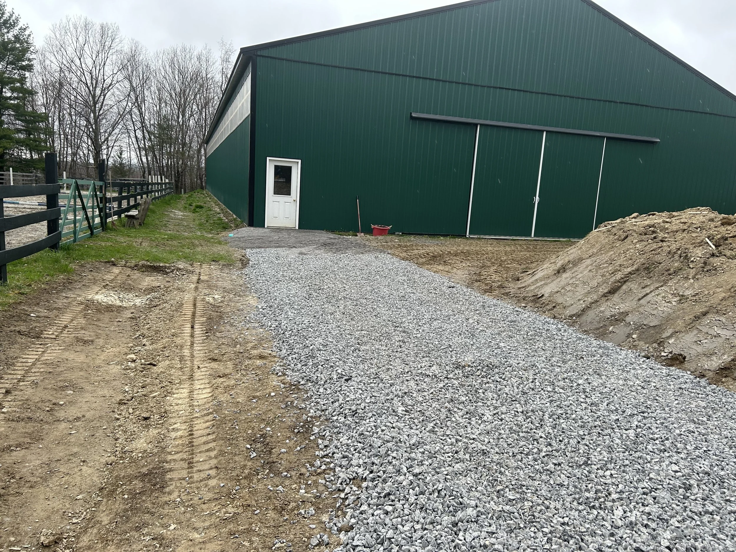 A gravel driveway in front of a large green metal barn with a white door and sliding doors, on a cloudy day with leafless trees in the background.