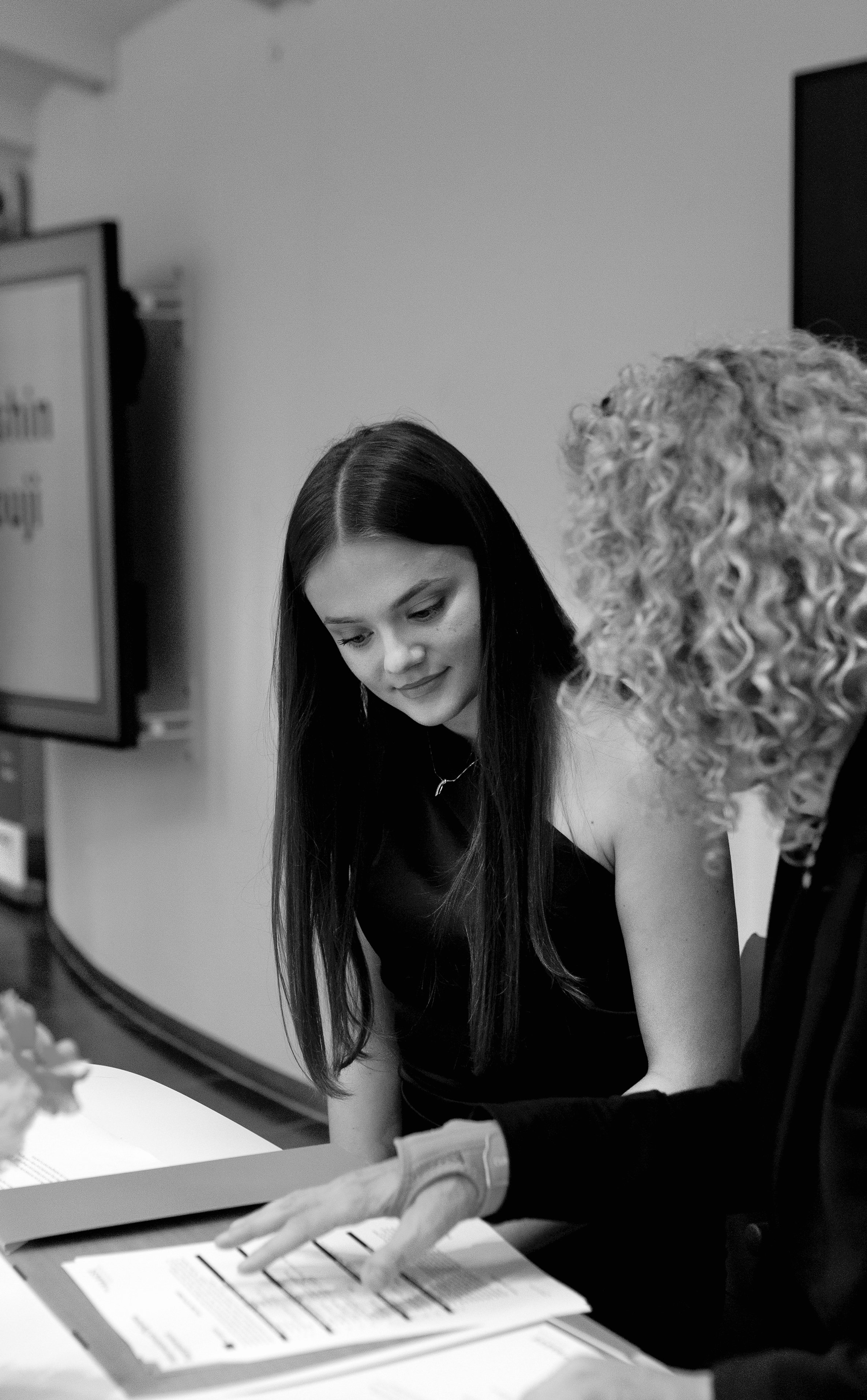 A young woman with long dark hair and a dark sleeveless dress looking at papers being shown by an older woman with curly blonde hair and glasses, in a professional setting.