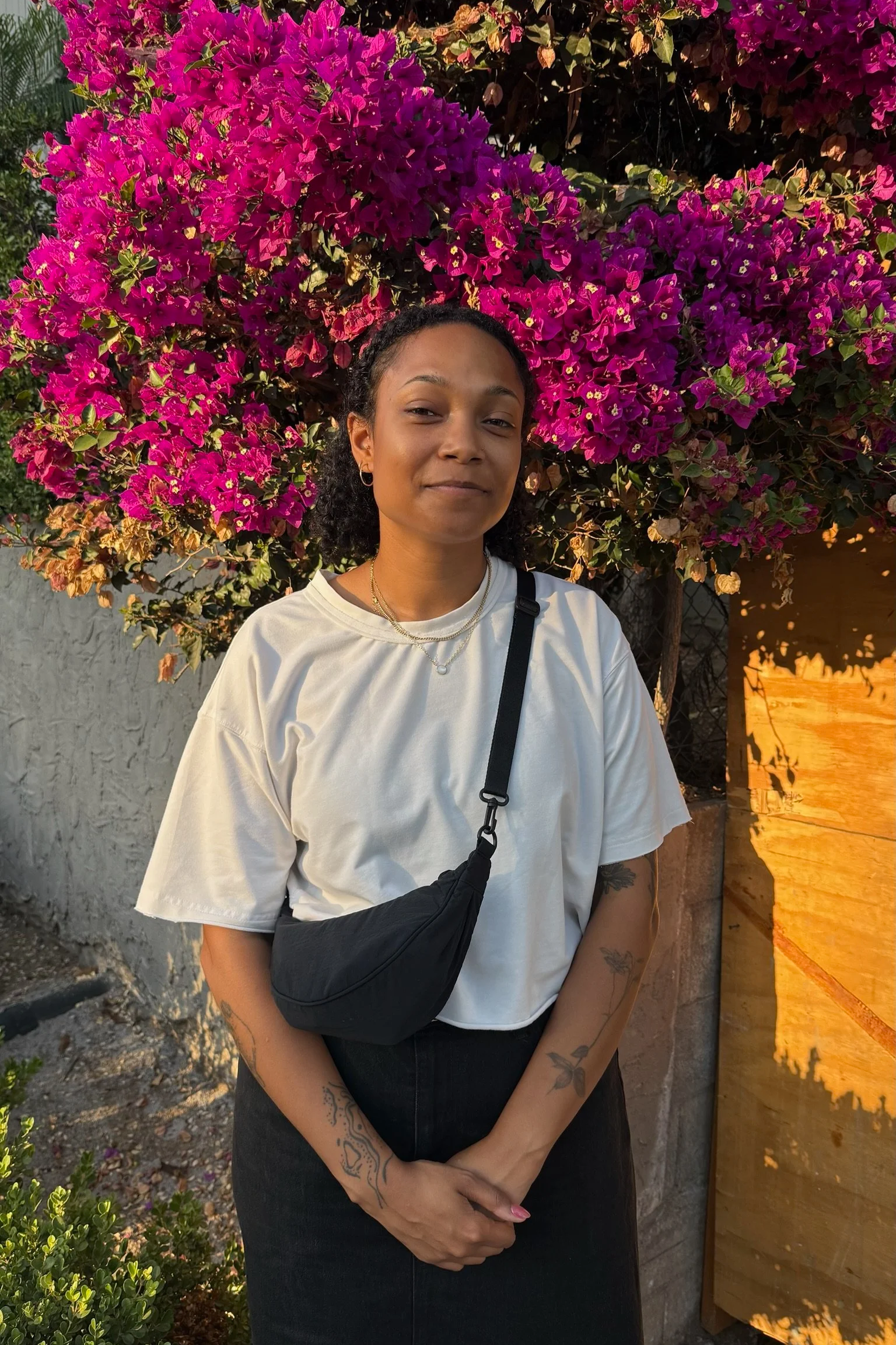 A young woman with curly dark hair standing in front of a large vibrant pink bougainvillea flower bush, wearing a white t-shirt, black crossbody bag, and tattoos on her arms, with a neutral expression.