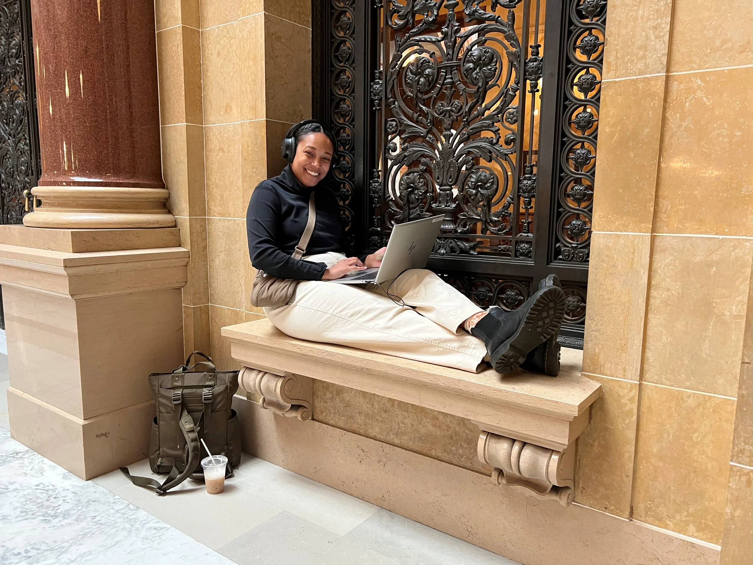 A woman with a black jacket and beige pants sitting on a stone bench inside a building, smiling, with a laptop on her lap wearing headphones. Next to her on the floor is a backpack, and on the ground is a beverage in a clear plastic cup with a straw.