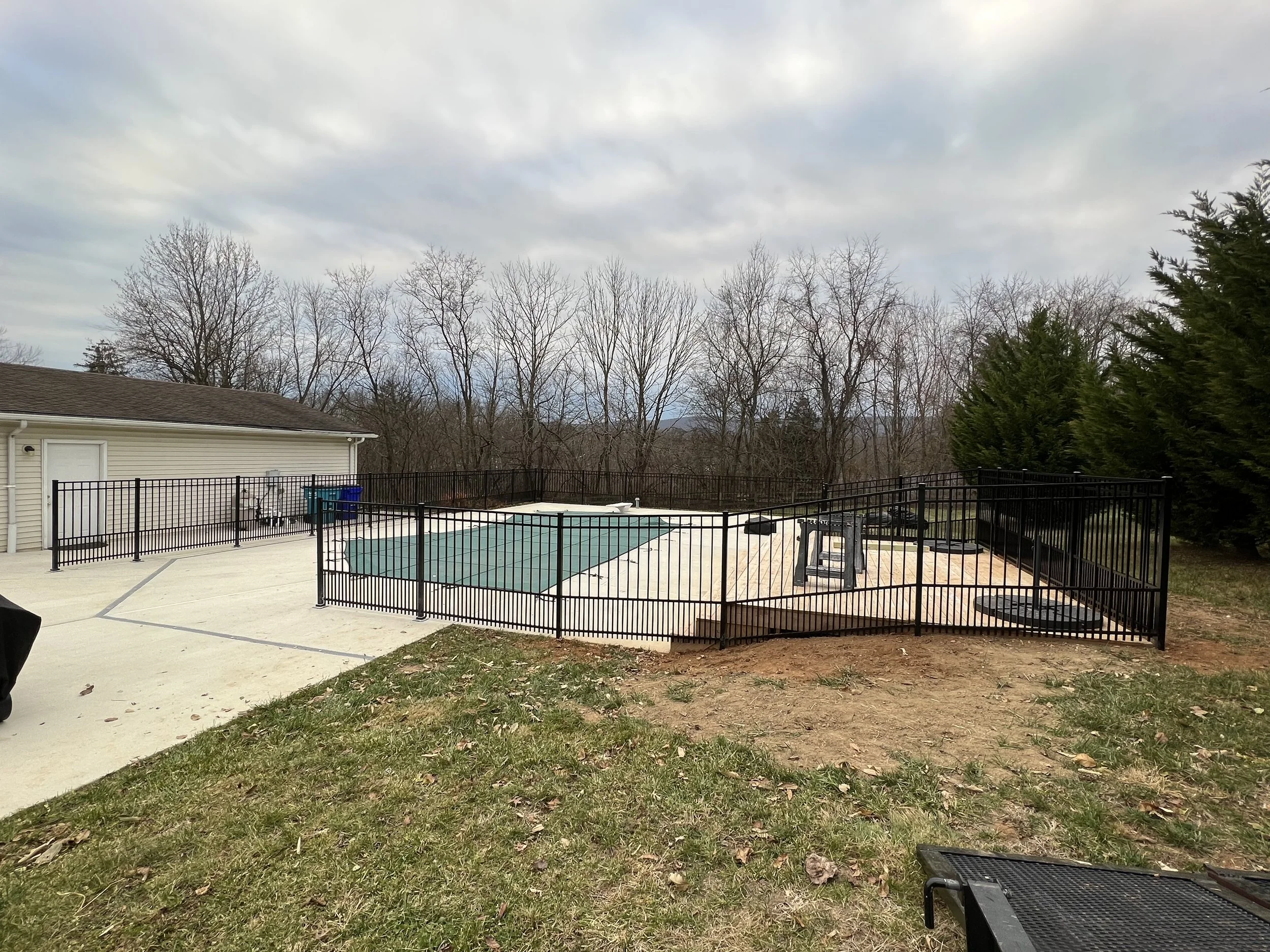 In-ground swimming pool enclosed by a black metal fence in a backyard, with a cloudy sky and leafless trees in the background.