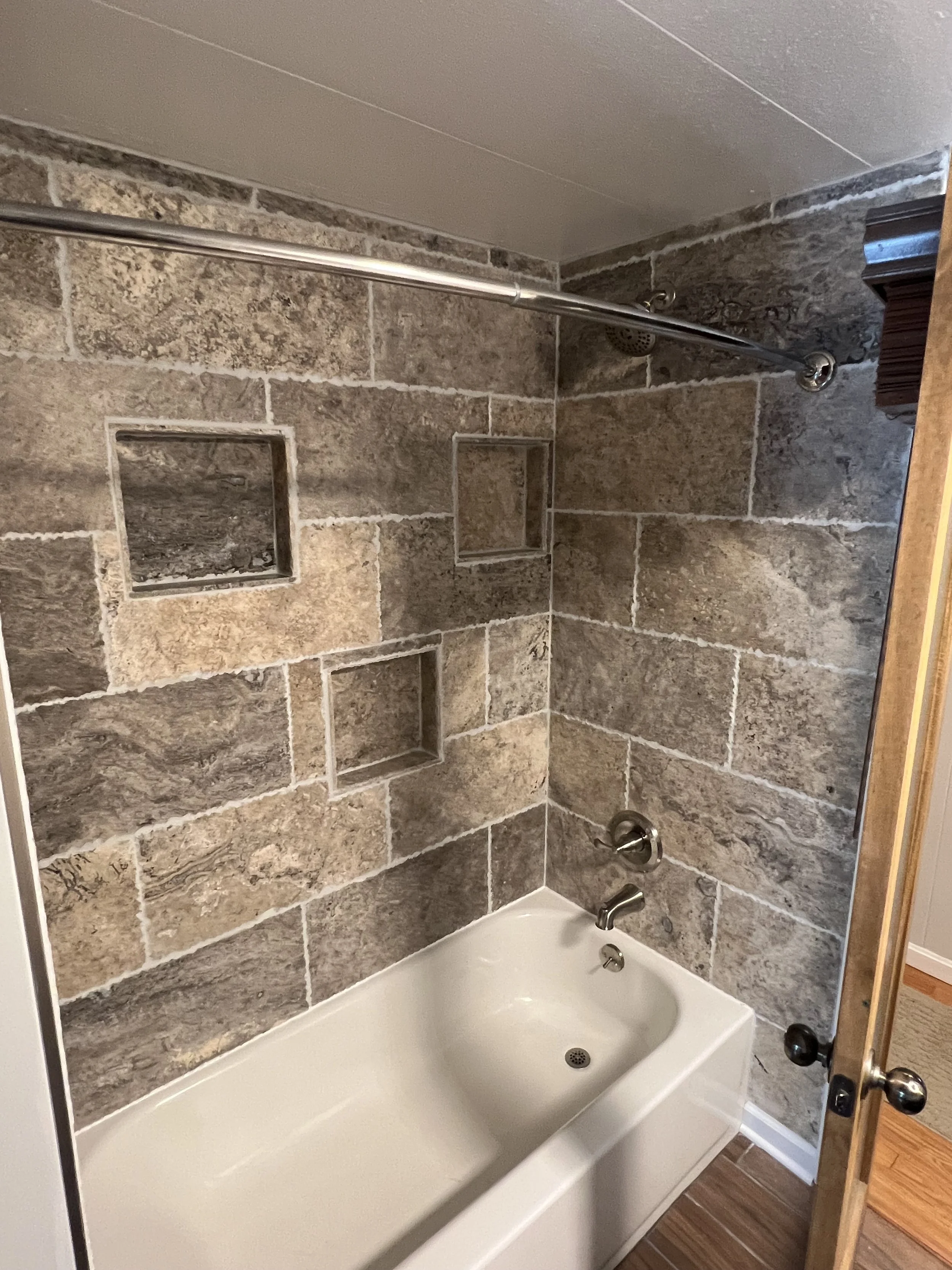 Bathroom with a bathtub, gray stone tiled wall with built-in niche shelves, showerhead, faucet, and a shower curtain rod.