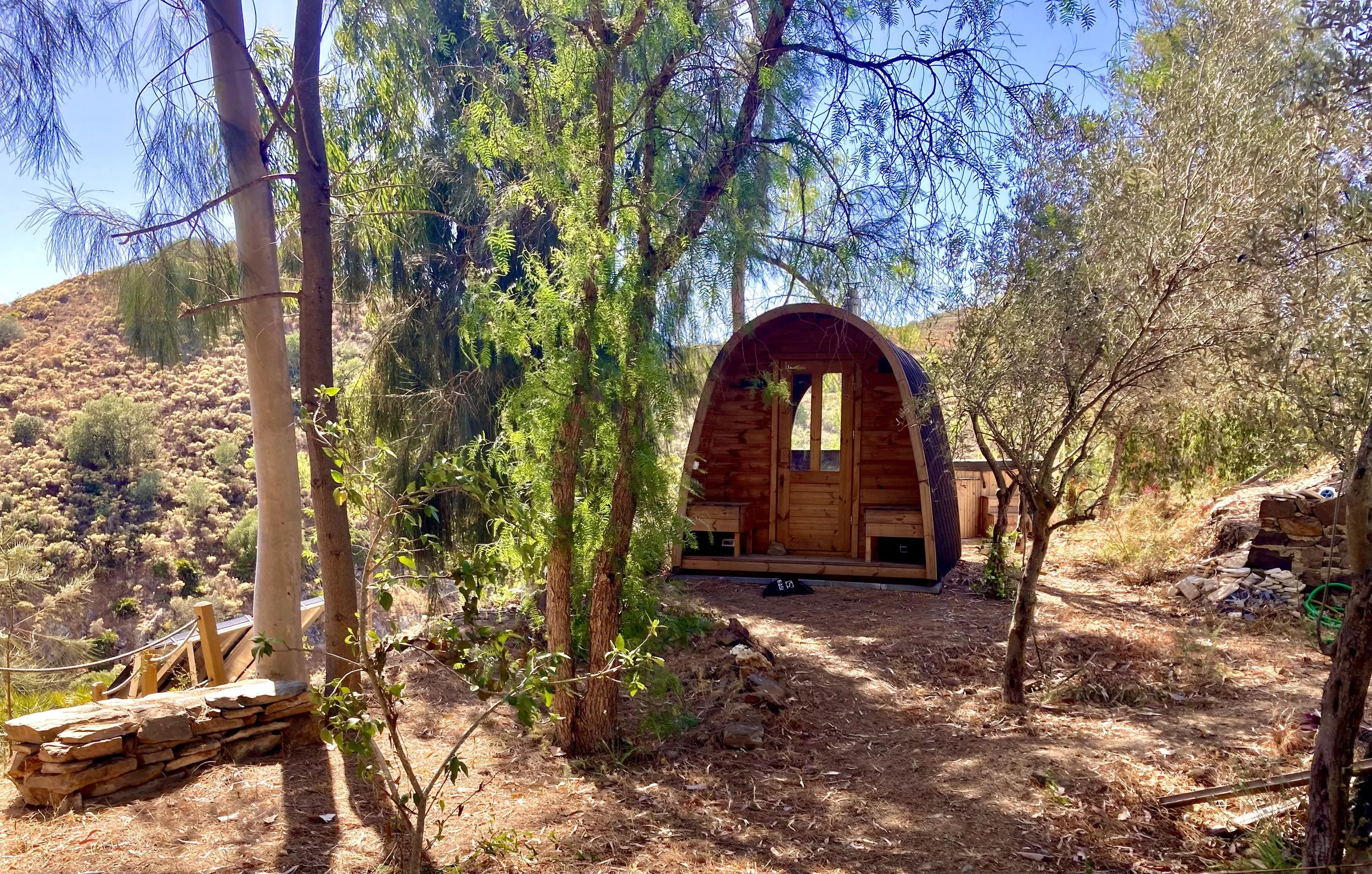 A small wooden dome-shaped cabin surrounded by trees on a hillside with dry terrain and a clear blue sky.