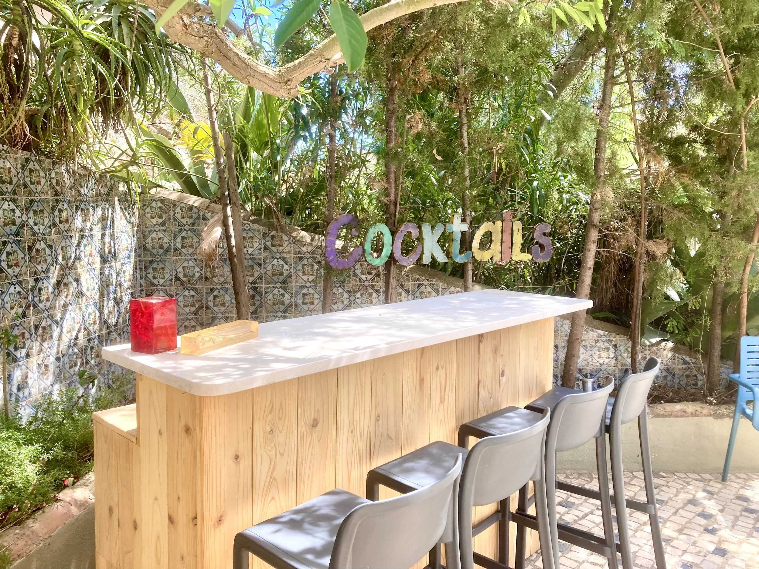 Colorful sign spelling 'Cocktails' behind a wooden bar counter with four gray barstools, set outdoors with lush green foliage and decorative tiles in the background.