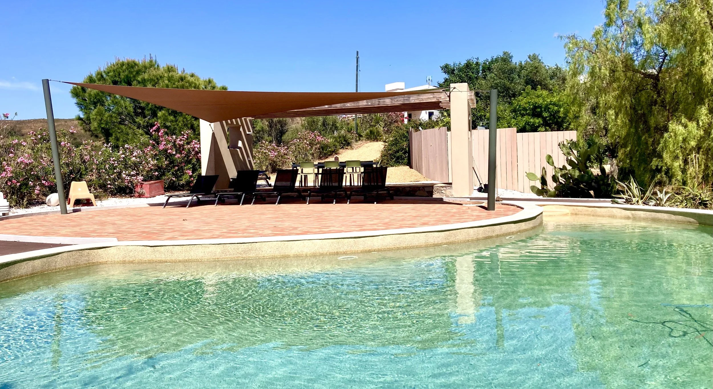 View of an outdoor swimming pool with clear water, surrounded by a patio with lounge chairs and a shaded seating area. There is a shade sail and a scenic background of trees, bushes, and a wooden fence under a clear blue sky.