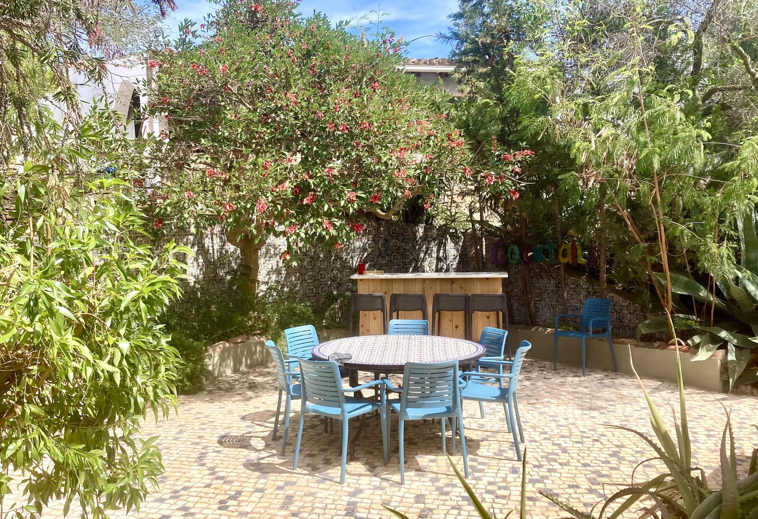 An outdoor patio area with a round table and blue chairs, surrounded by lush green plants and trees, with a small bar in the background, and a colorful "cocktail" sign on the fence.