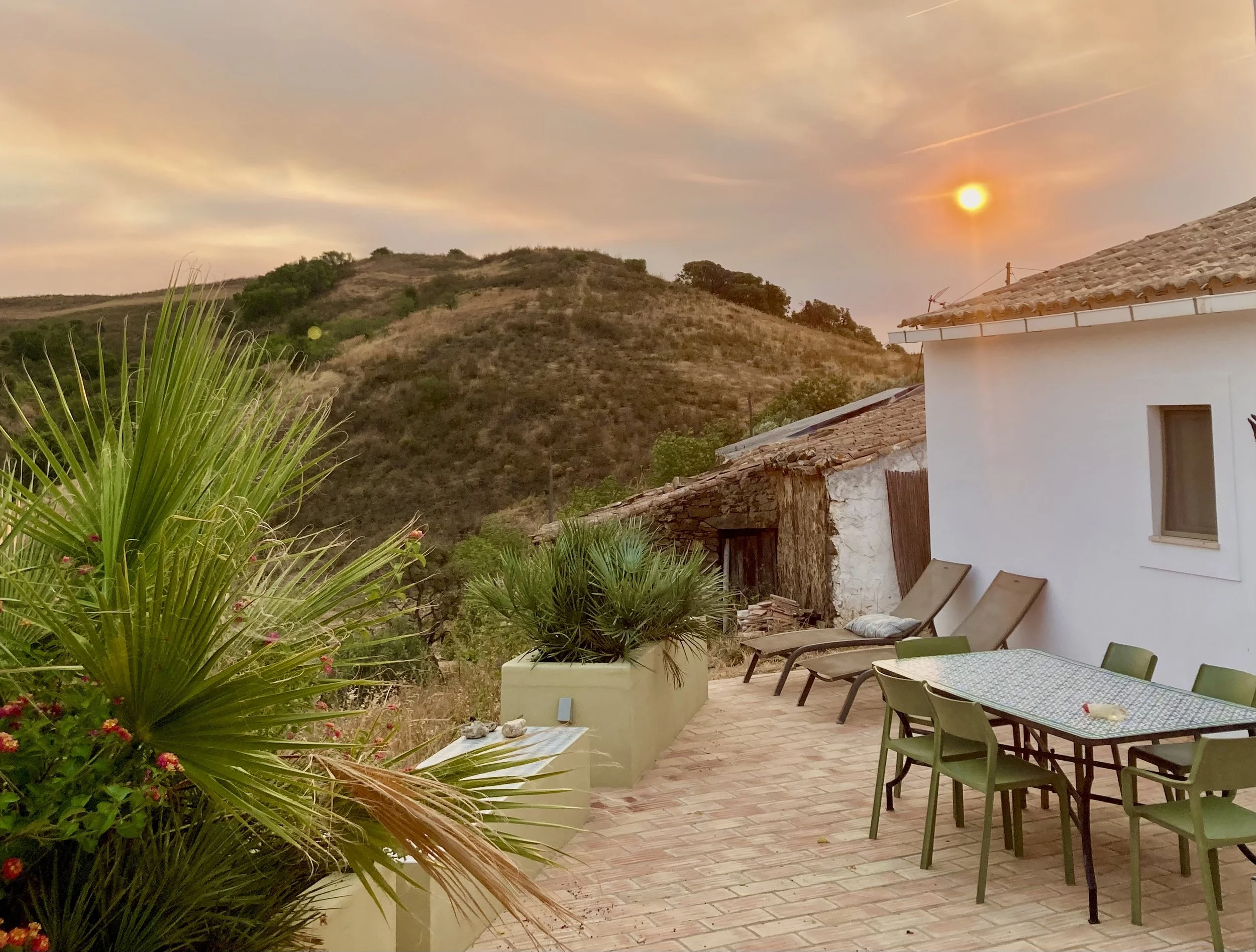 Sunset over hills seen from a patio with green chairs, a rectangular glass-topped table, lounge chairs, potted plants, and a white house with a tiled roof