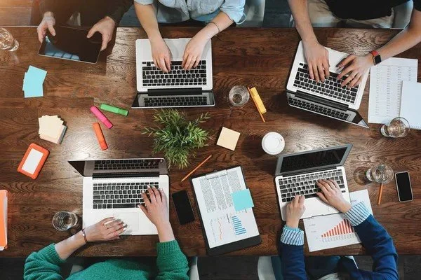 Top-down view of people working on laptops at a wooden table with office supplies, notebooks, tablets, and a plant.