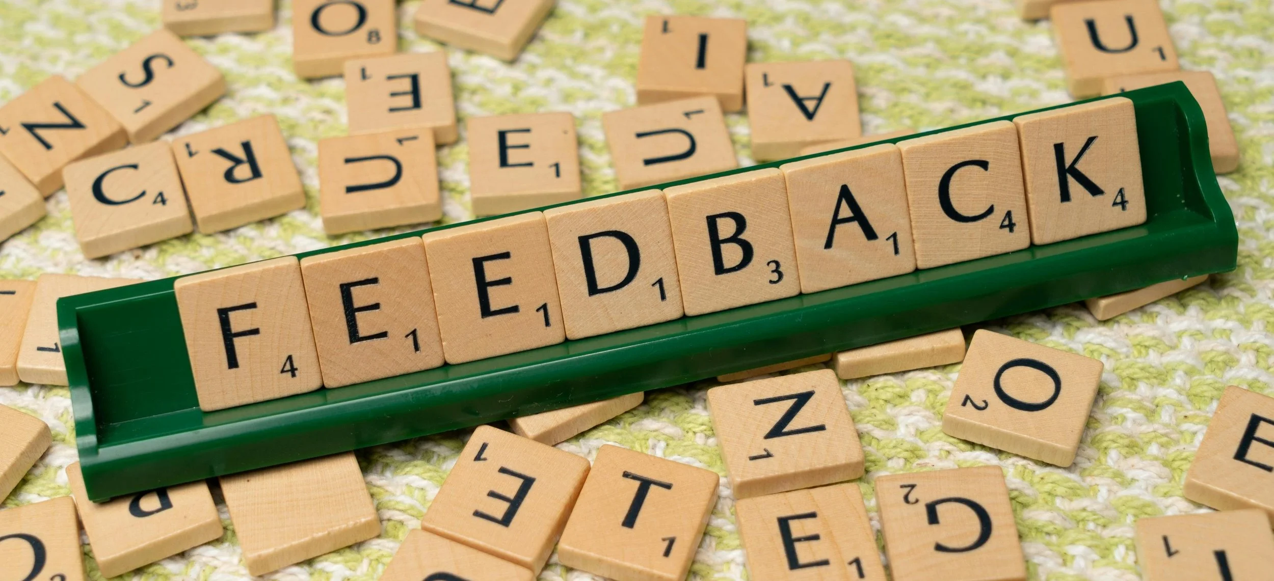 Scrabble tiles spelling the word 'FEEDBACK' on a green tray, scattered on a patterned surface.