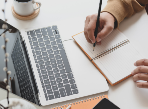 Person writing in a spiral notebook with a black pen, beside a silver laptop on a white desk.