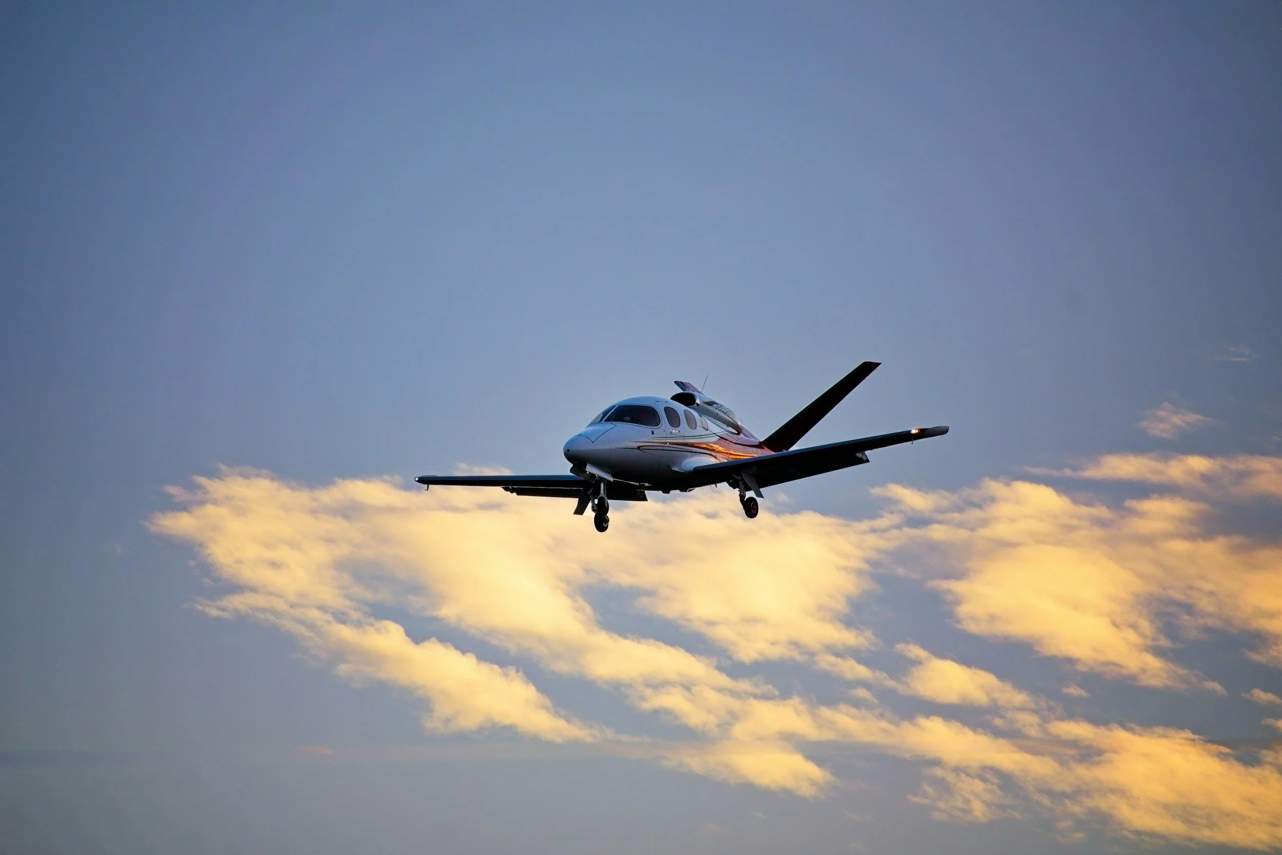 A small private jet flying in the sky at sunset with clouds below.