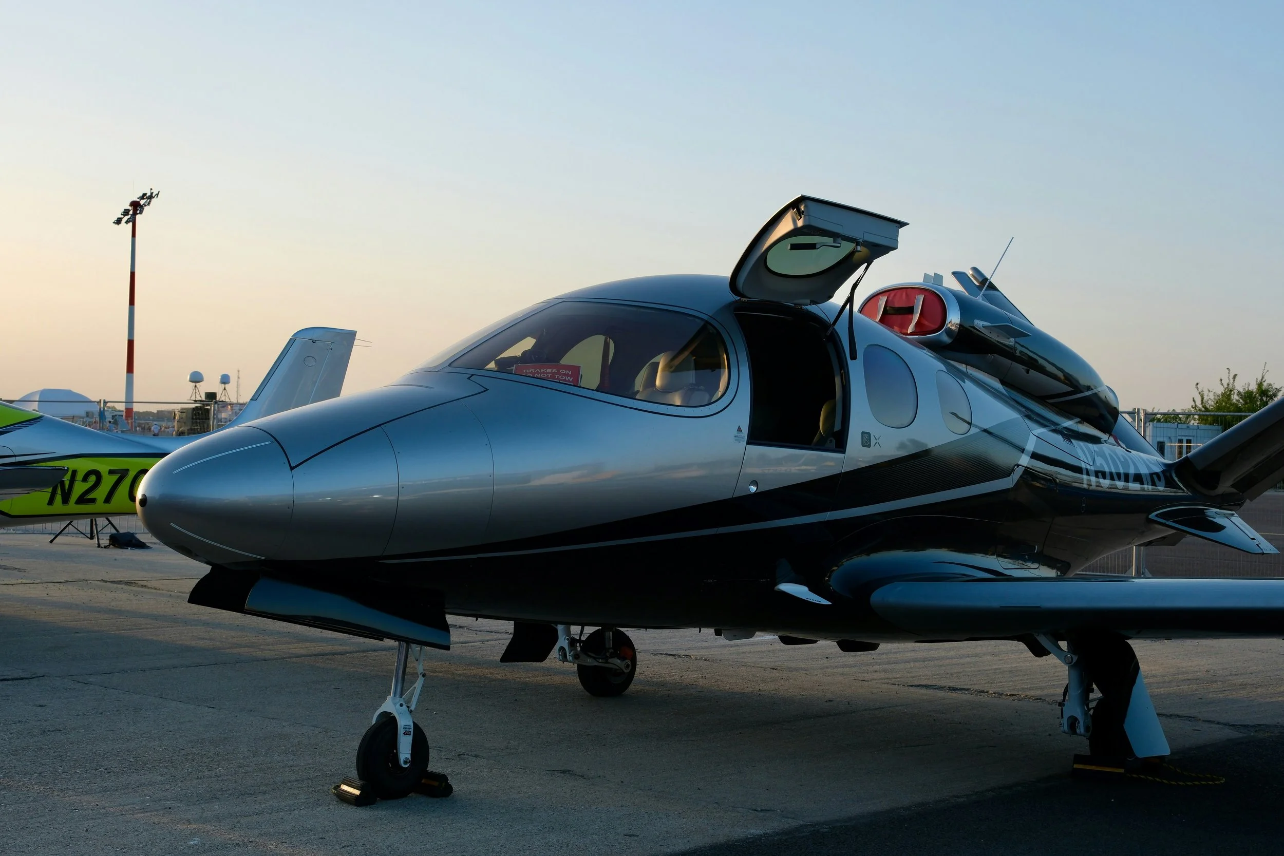 A sleek, futuristic aircraft parked on a tarmac at an airport during sunset, with a small engine mounted on top and its canopy open.