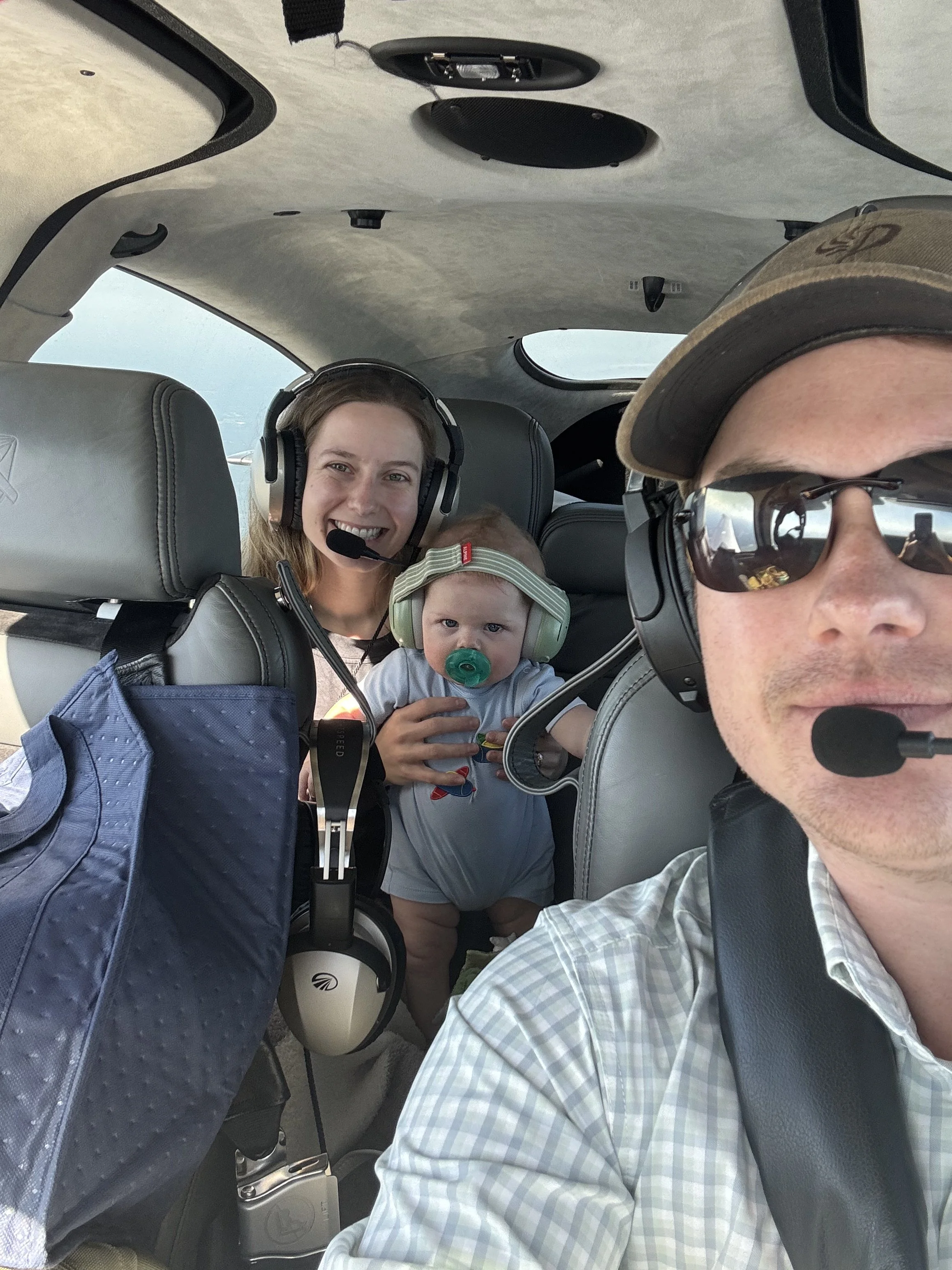 Family flying in a small airplane, with two adults, a young woman and a man, and a toddler, all wearing headsets and sitting inside the plane.