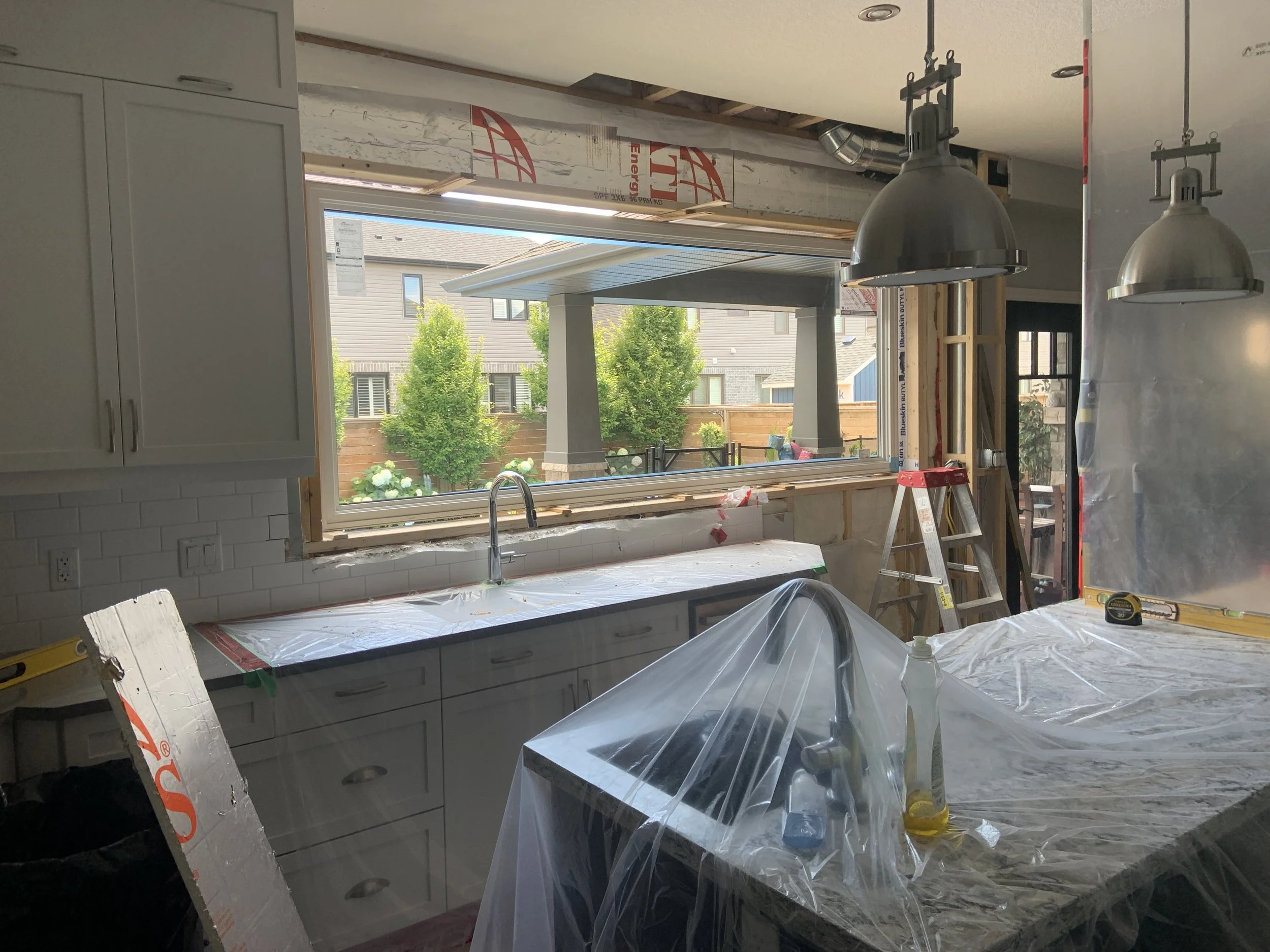 Indoor kitchen under construction with large window showing backyard, cabinetry, lighting fixtures, construction materials, and tools visible.